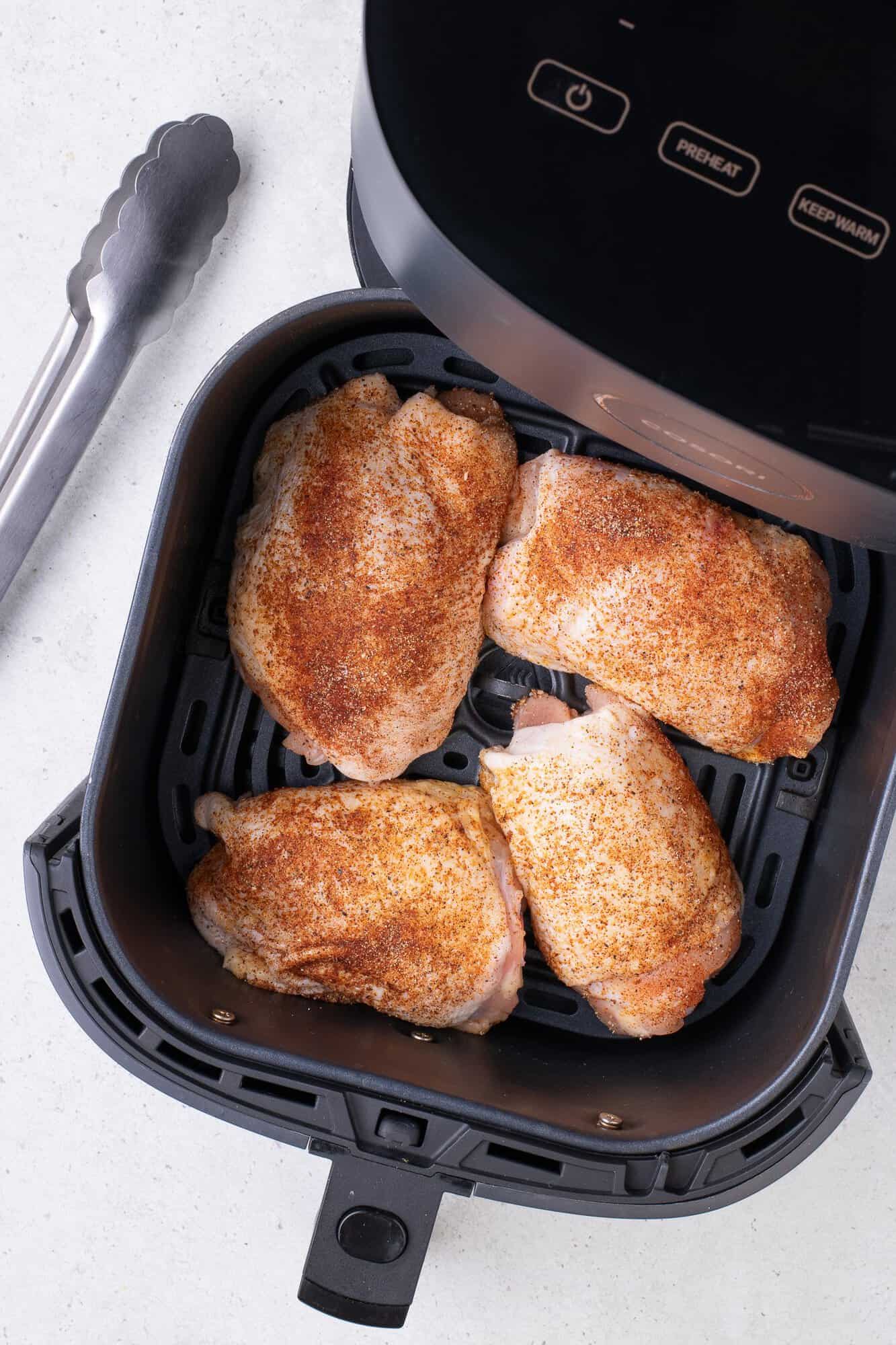 Chicken thighs seasoned with spices are placed inside an air fryer basket, ready for cooking. Tongs are positioned beside the air fryer.