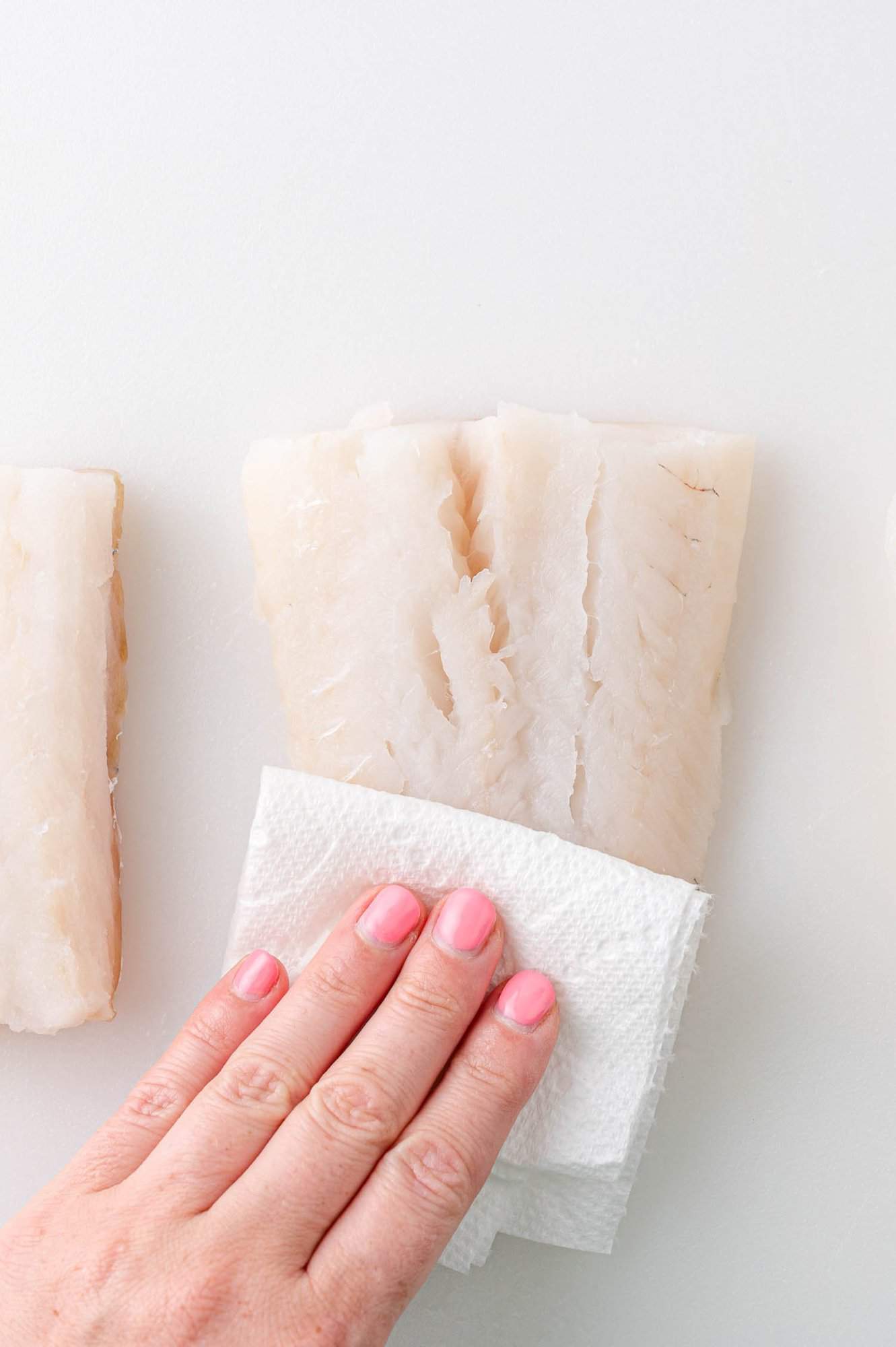 A hand pats dry a piece of raw fish with a paper towel on a white surface.