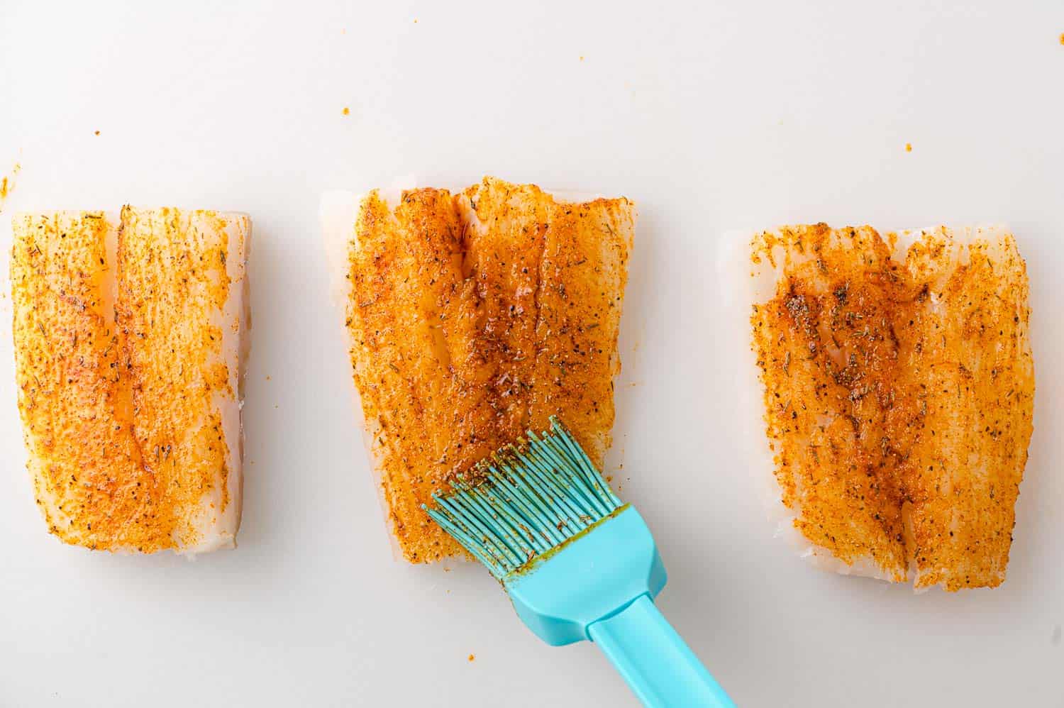 Cod fillets on a white surface being brushed with spices using a blue silicone brush.