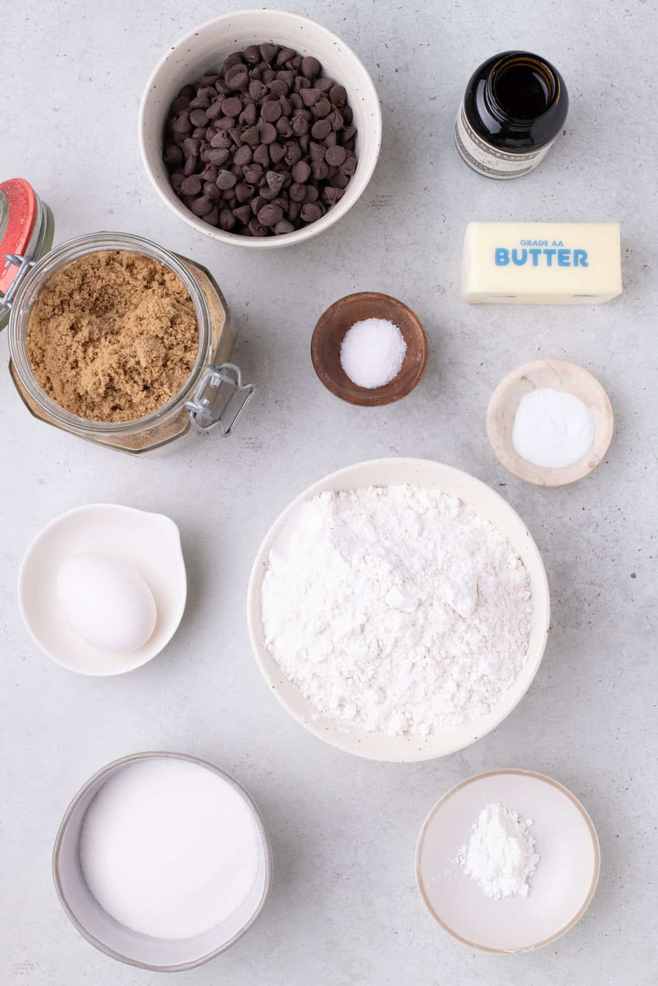 Ingredients for baking: chocolate chips, vanilla extract, butter, brown sugar, salt, baking soda, flour, egg, white sugar, and baking powder are arranged on a countertop.