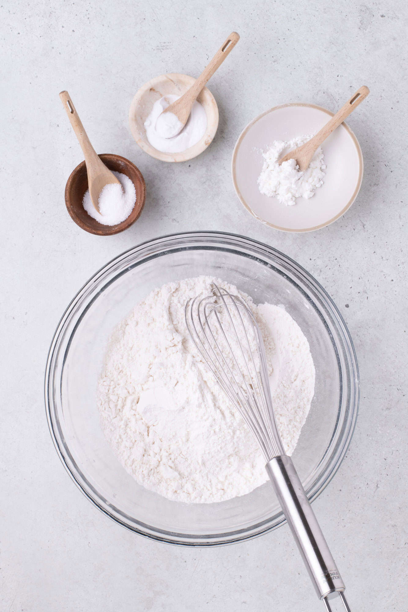 Bowl of flour with a whisk, surrounded by small bowls containing salt, baking powder, and baking soda on a light surface.