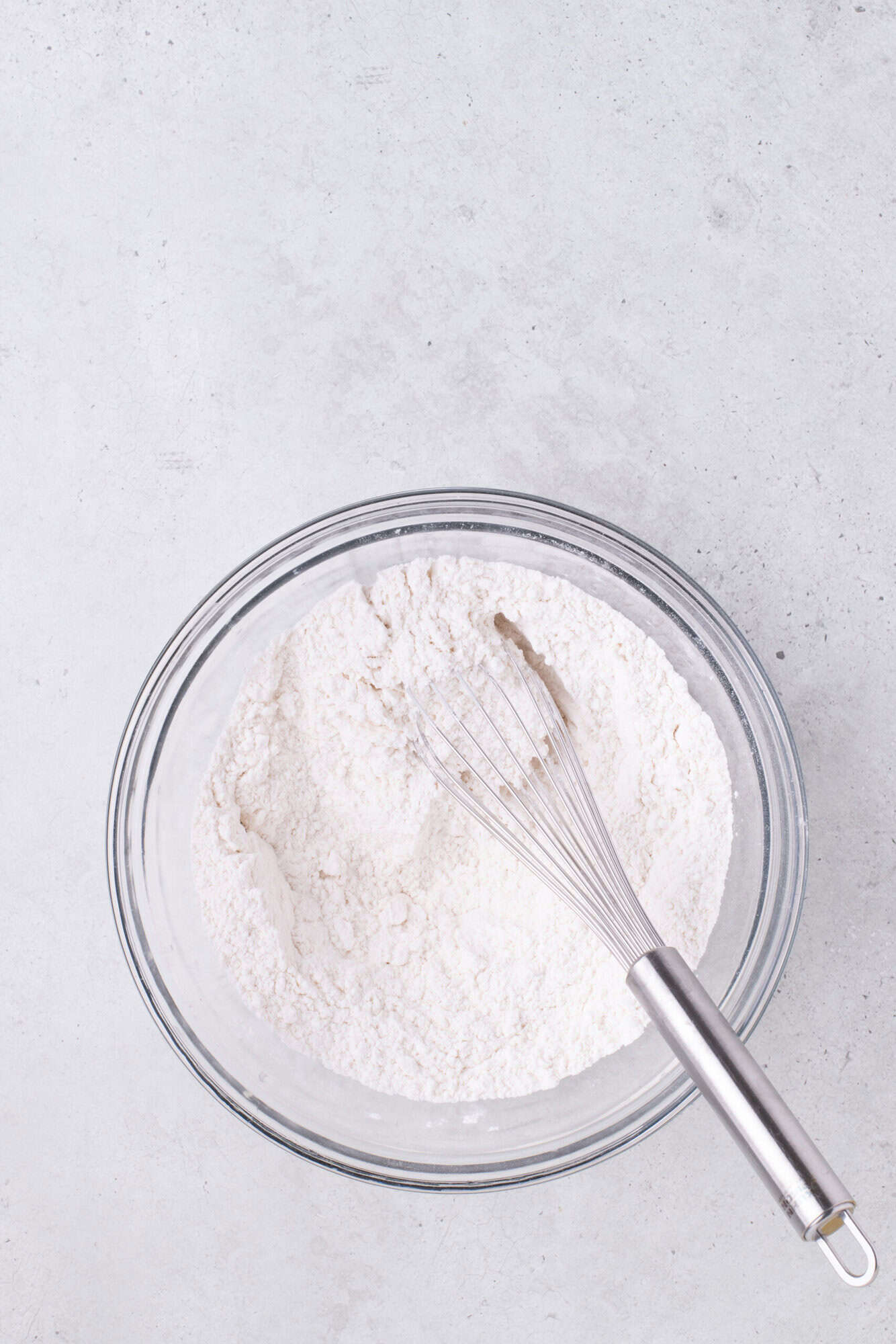 A glass bowl filled with flour and a metal whisk on a light gray surface.