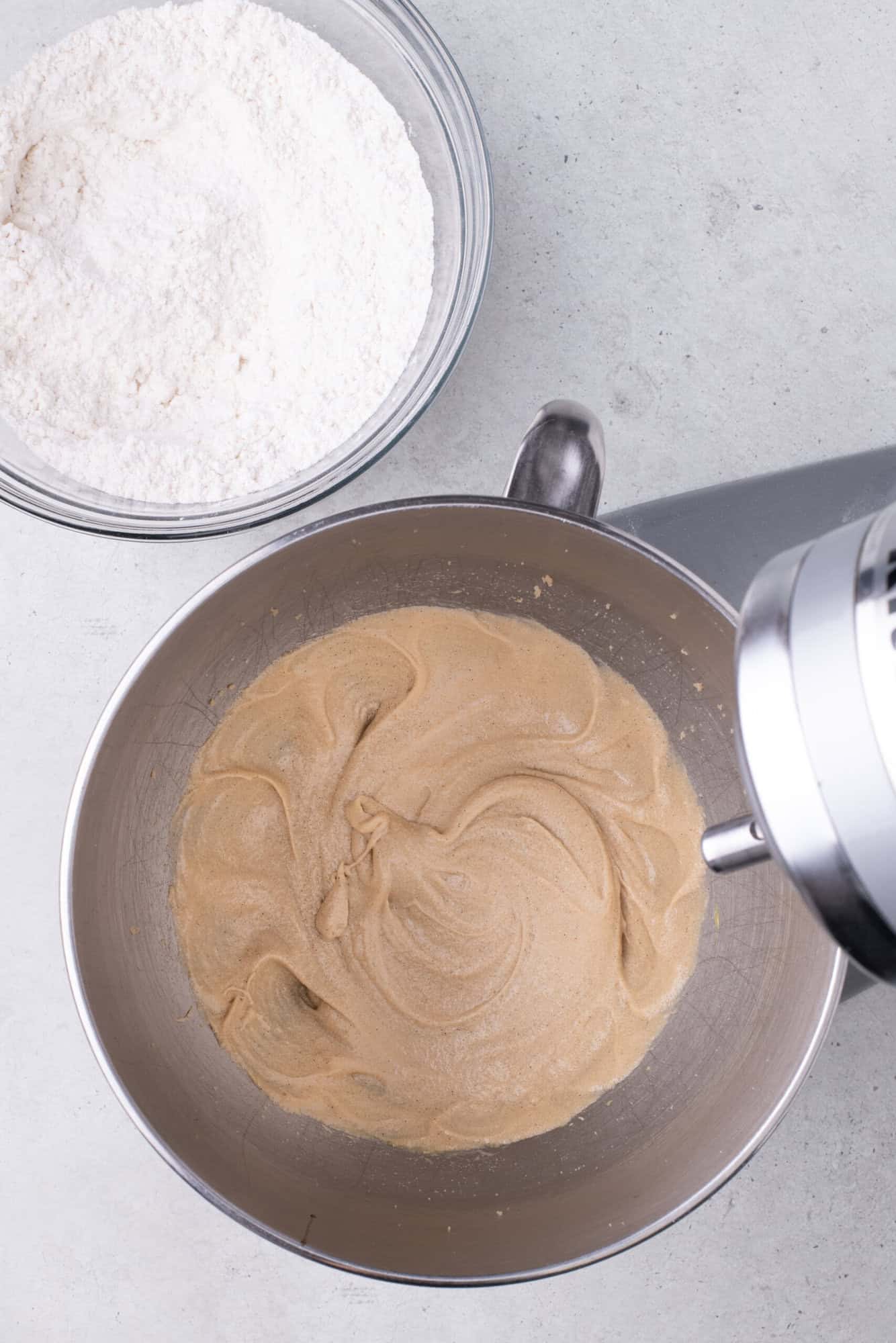 A mixing bowl with brown batter is attached to a stand mixer. A glass bowl with white flour is nearby on a gray surface.