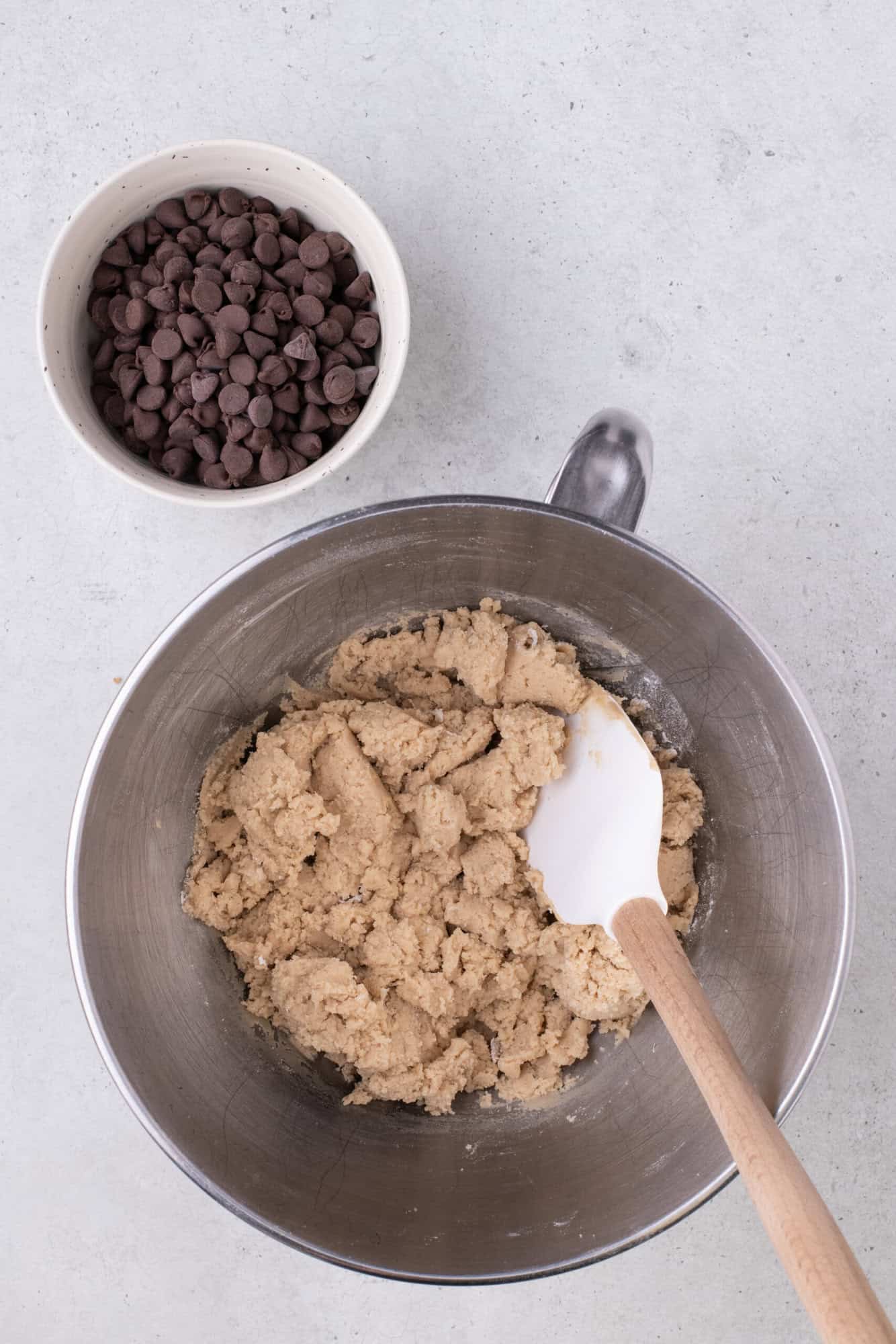 Mixing bowl with cookie dough and a spatula, next to a small bowl of chocolate chips on a light surface.