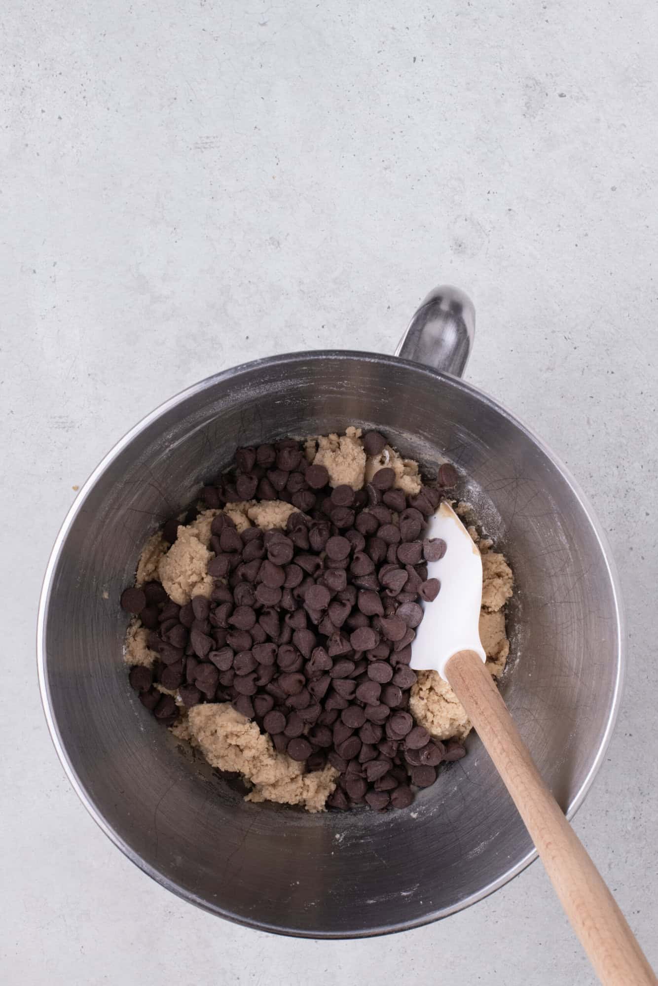 Bowl of cookie dough with chocolate chips and a white spatula on a gray surface.