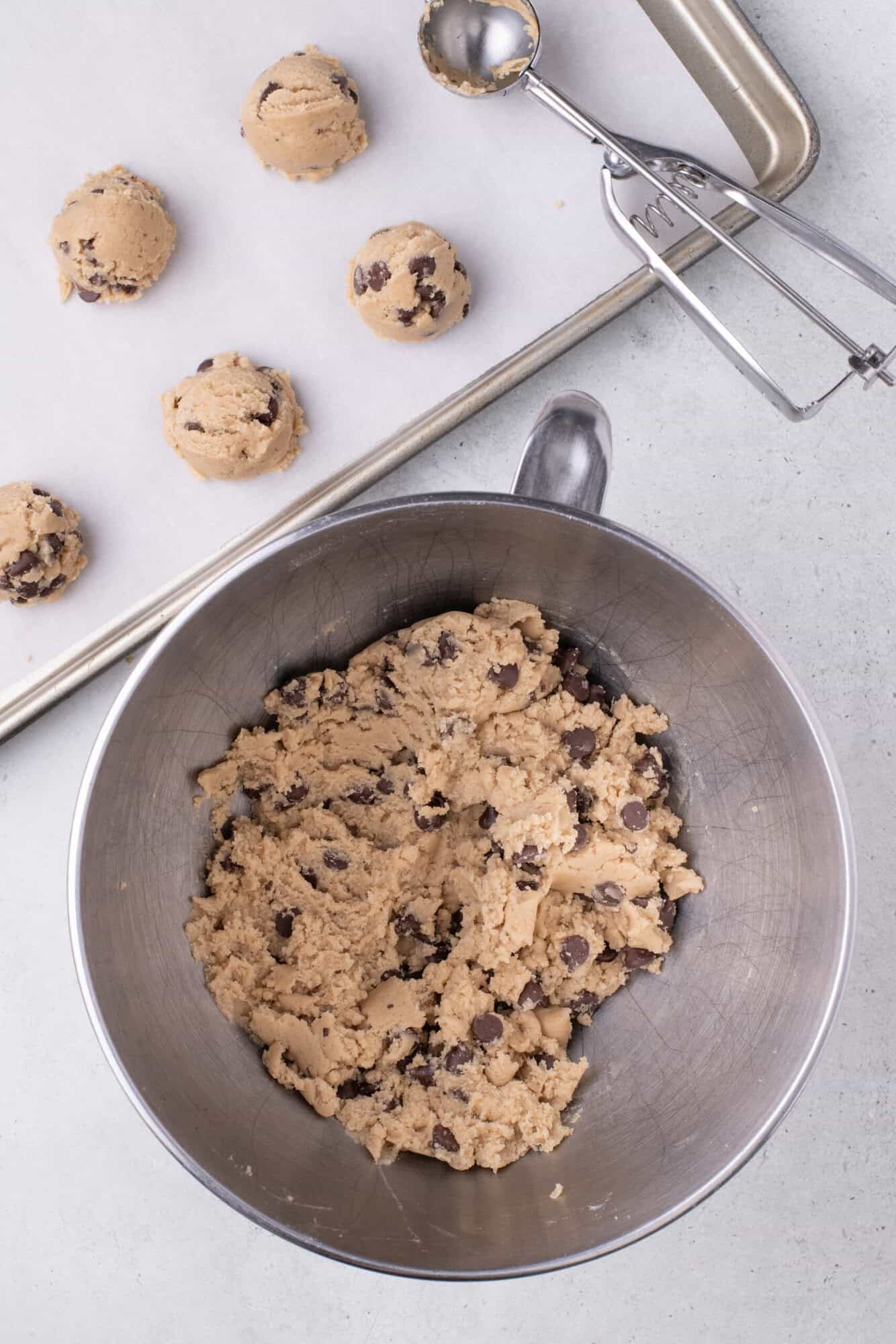 Chocolate chip cookie dough in a mixing bowl with several dough scoops on a baking tray and a cookie scoop nearby.