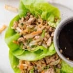 Ground meat and vegetables wrapped in lettuce leaves, served with a small dish of dark dipping sauce on a white plate.
