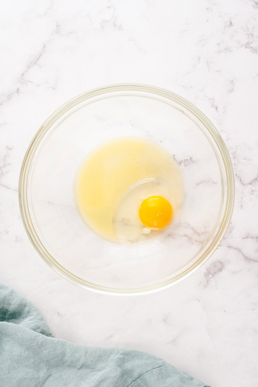 A cracked egg and sugar in a clear glass bowl on a white marble surface, with a light blue cloth nearby.