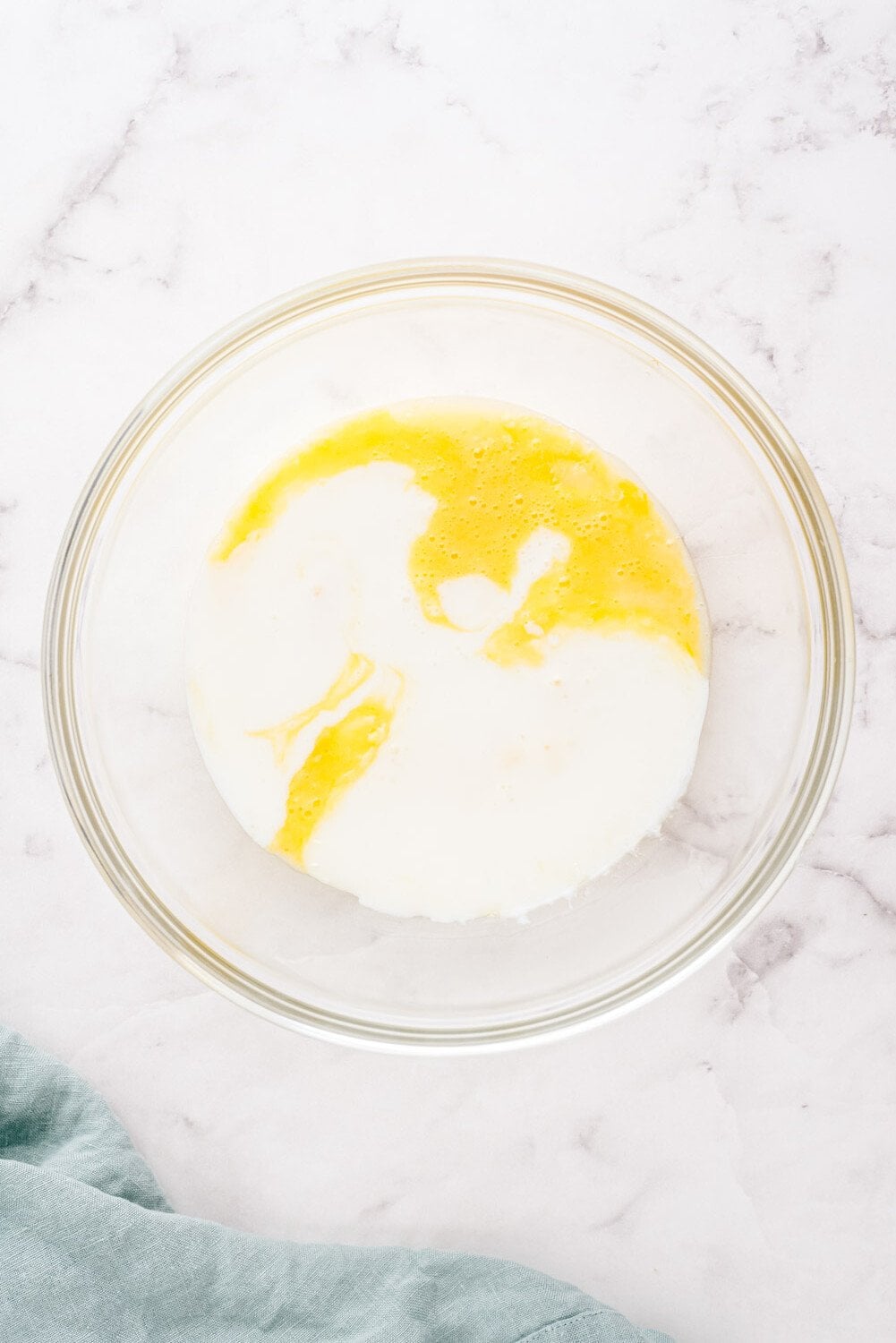 A glass bowl containing a mixture of milk and beaten eggs on a marble countertop, next to a blue cloth.