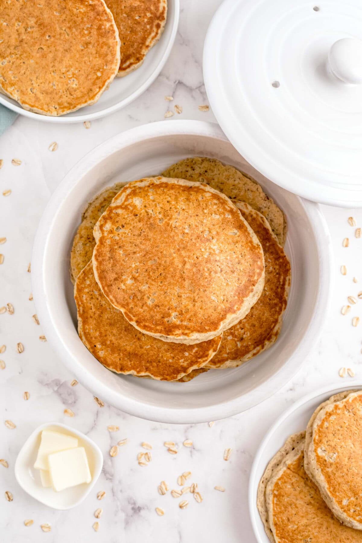 A top view of a white bowl filled with oat pancakes on a marble surface, surrounded by scattered oats.