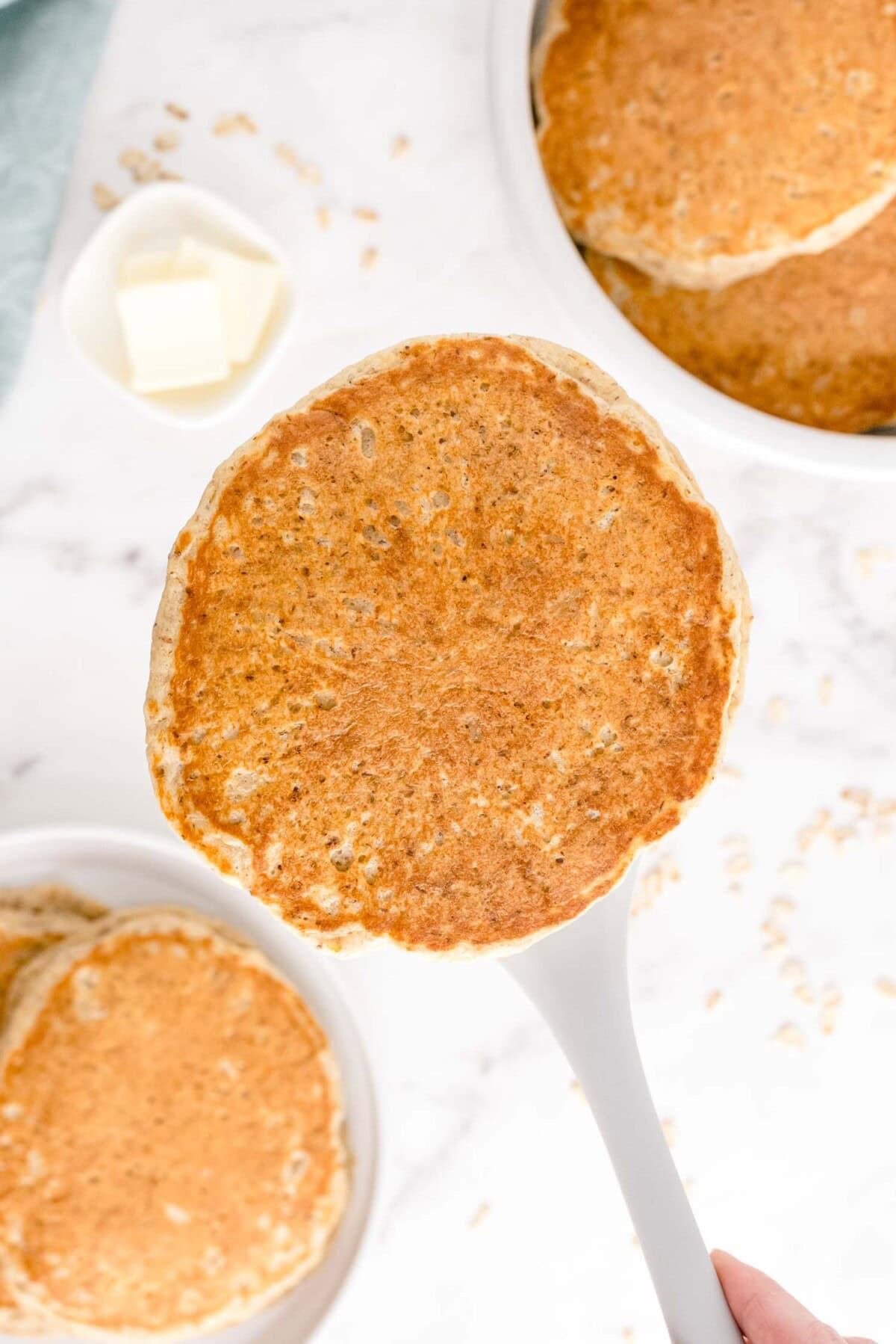 A spatula holding a golden-brown pancake over a plate of pancakes on a marble surface, with a dish of butter in the background.