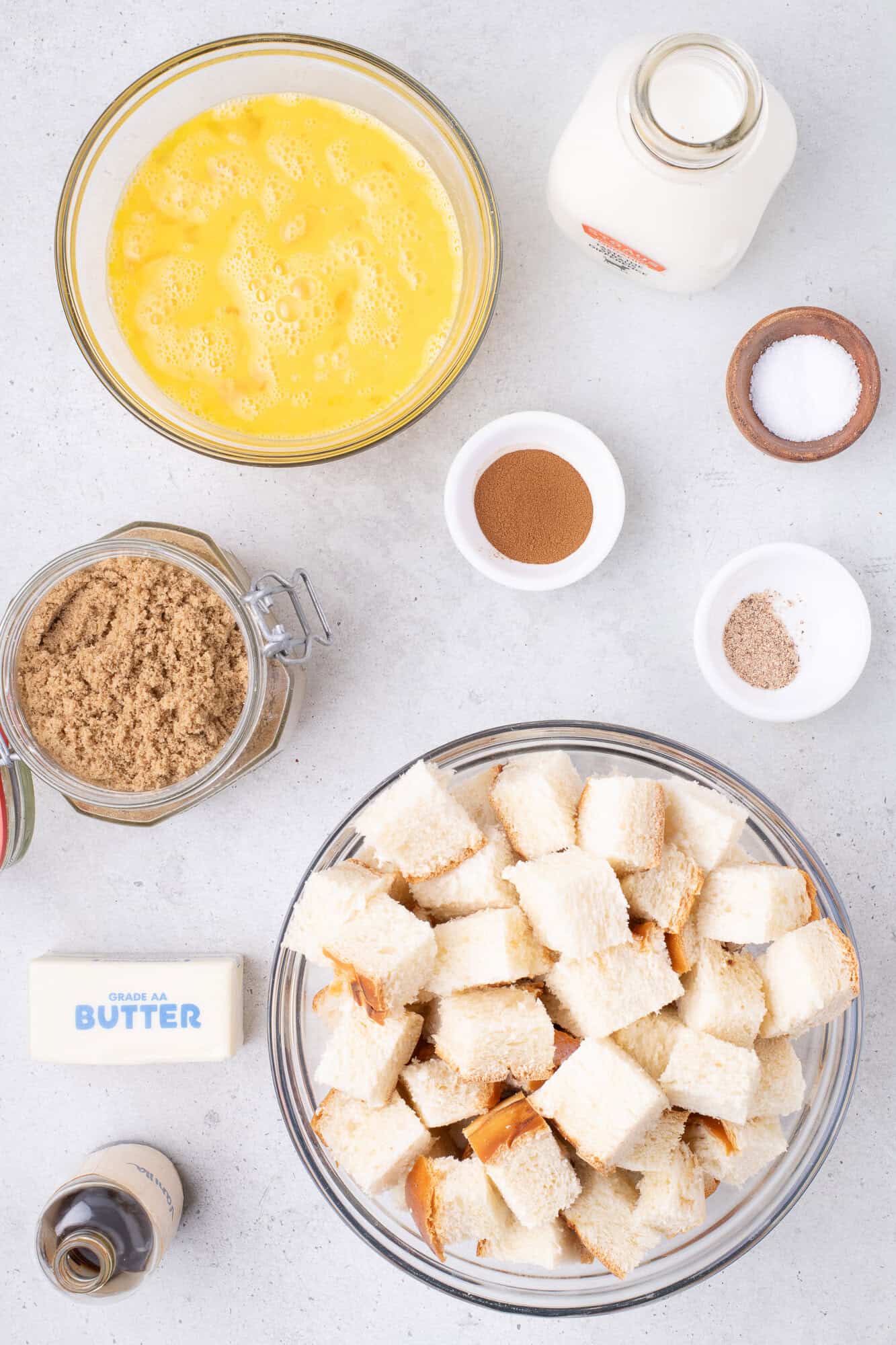 Ingredients for recipe are laid out: cubed bread, beaten eggs, milk, brown sugar, butter, cinnamon, and salt, arranged on a light-colored surface.