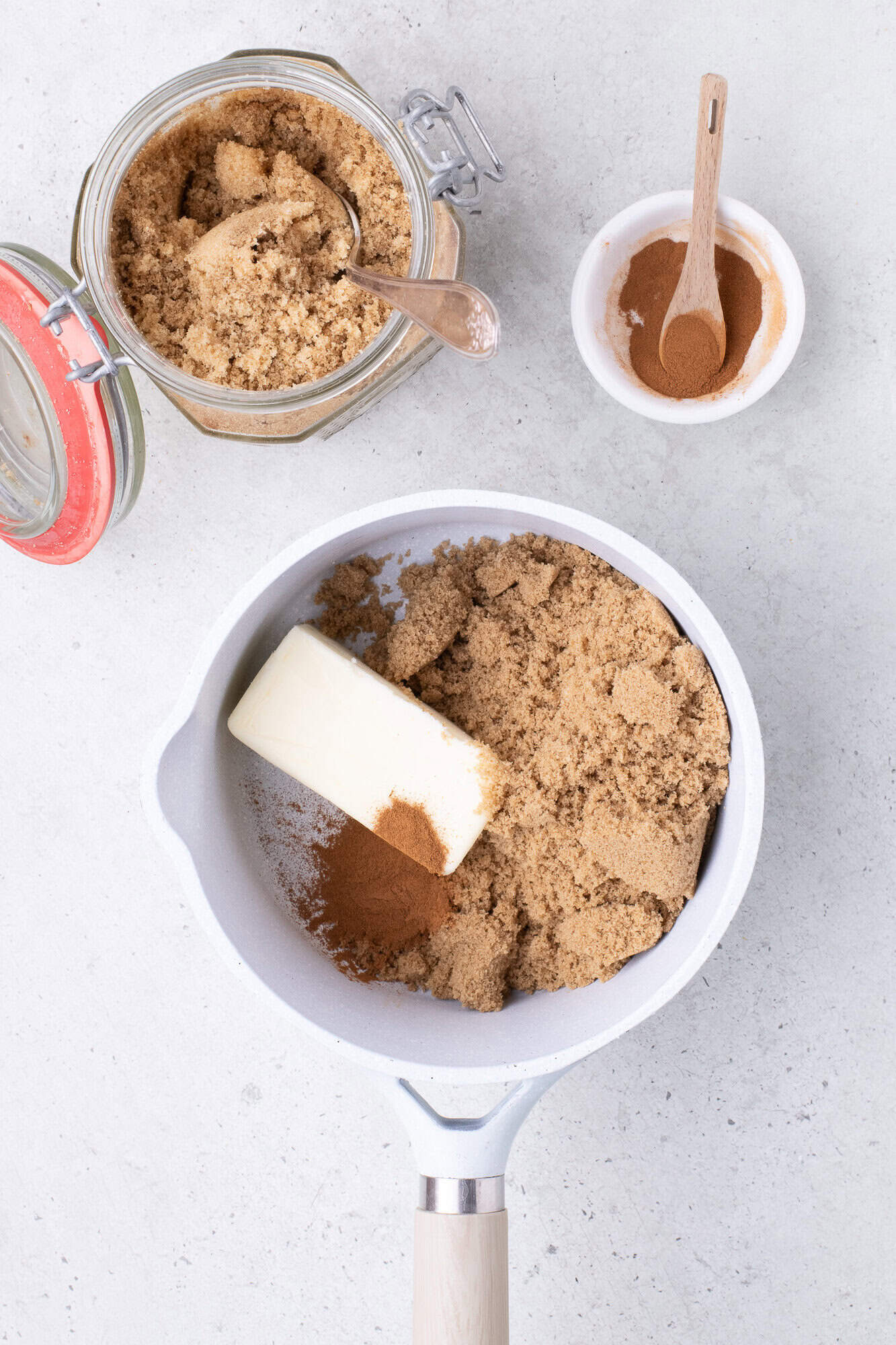 Butter, brown sugar, and cinnamon in a white saucepan, with extra sugar in a jar and cinnamon in a small bowl nearby on a light surface.