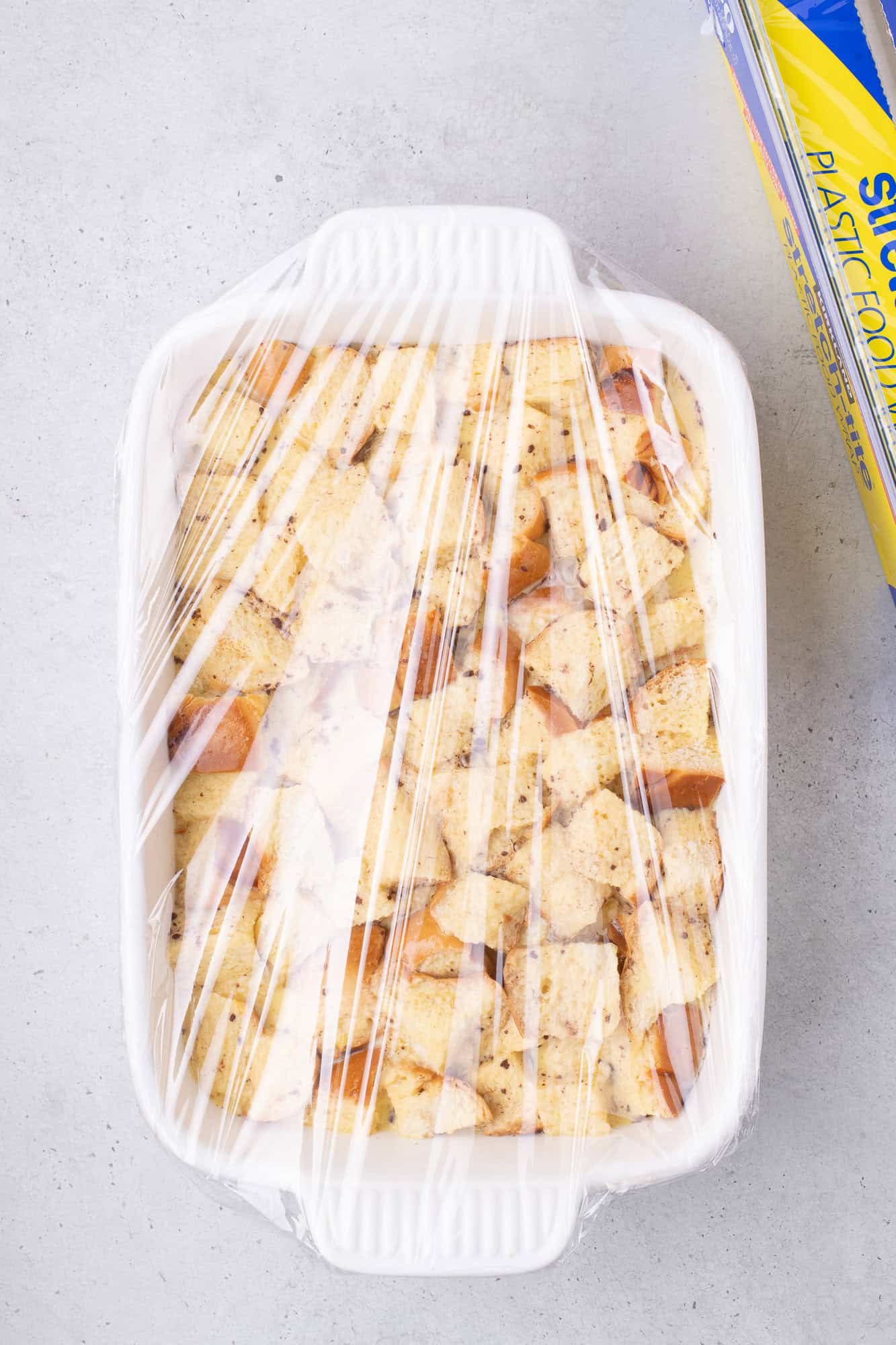 A white casserole dish filled with cubed bread is wrapped in plastic wrap. A box of plastic wrap is nearby on a gray surface.