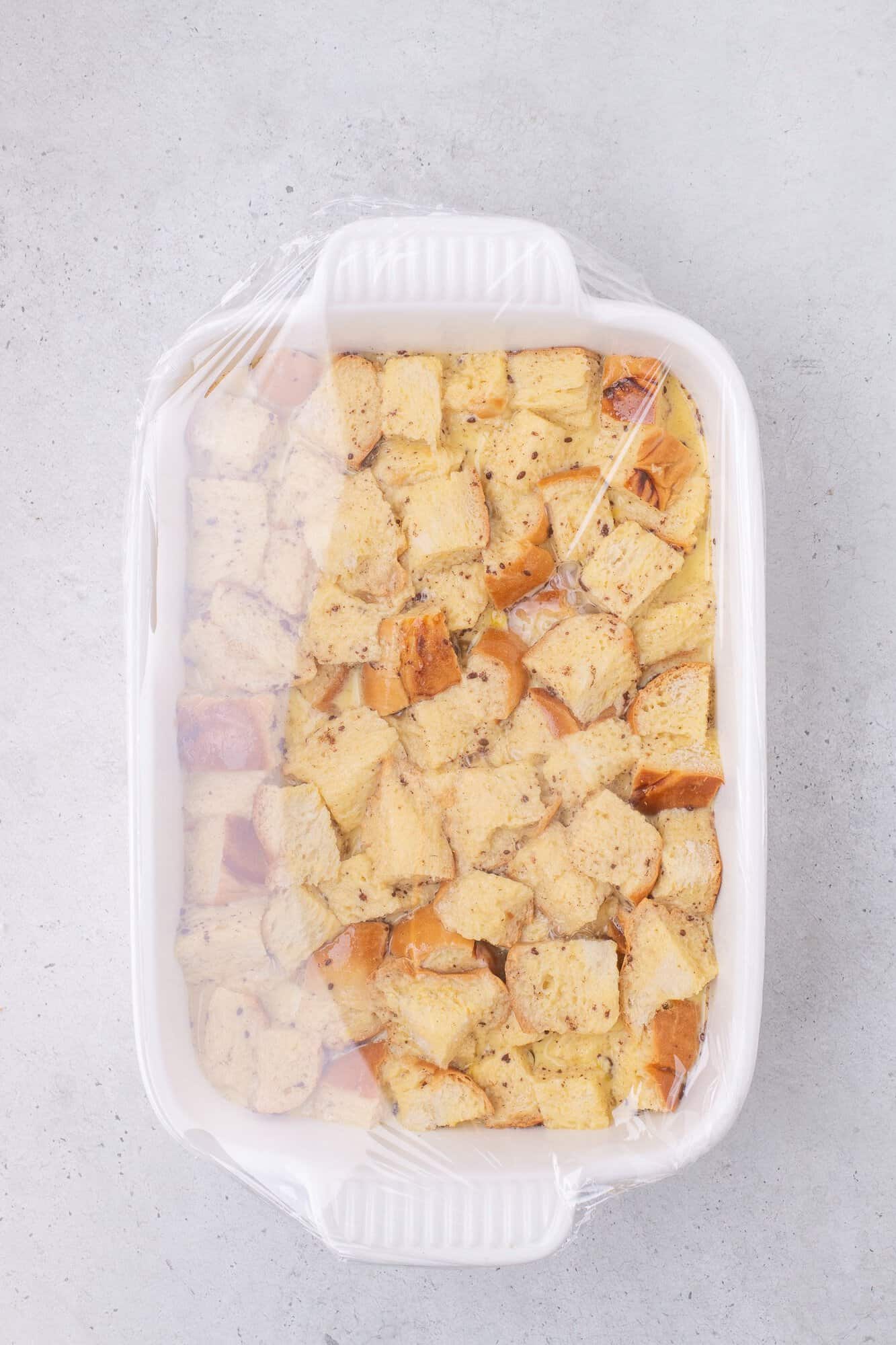 A white baking dish filled with cubed bread covered with plastic wrap, ready for baking.