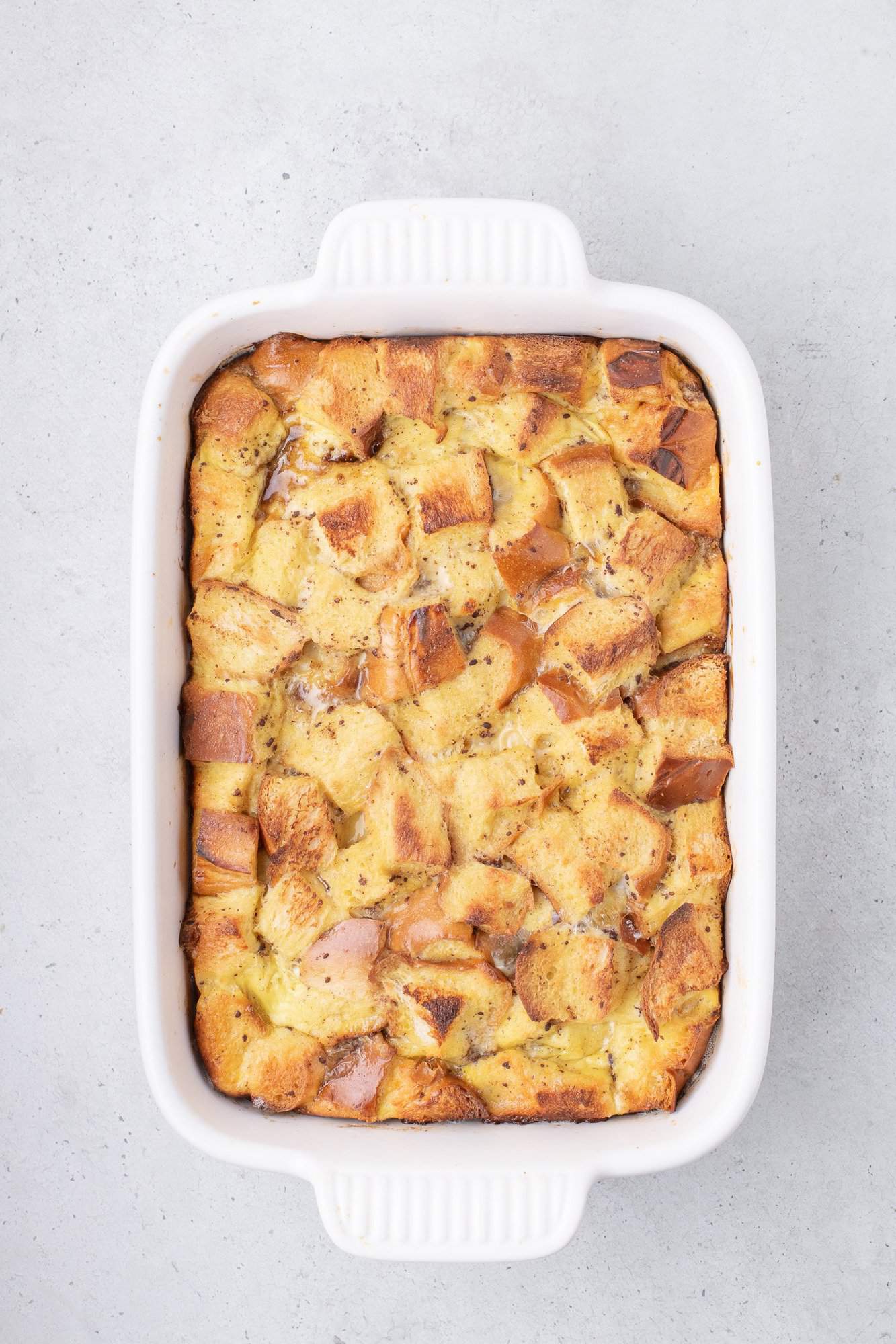 A baked french toast in a white rectangular dish, featuring golden brown bread cubes in a creamy mixture, on a light gray surface.