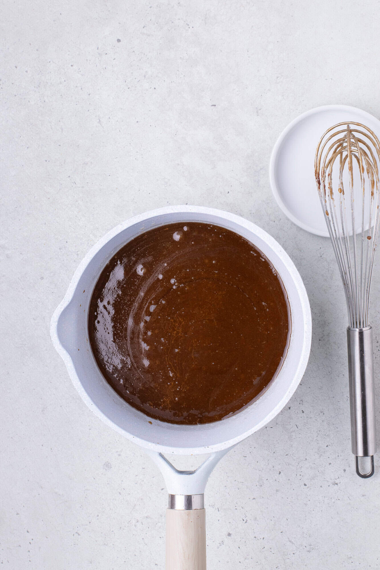 A white pot containing chocolate sauce sits on a light countertop, next to a whisk and an empty white bowl.