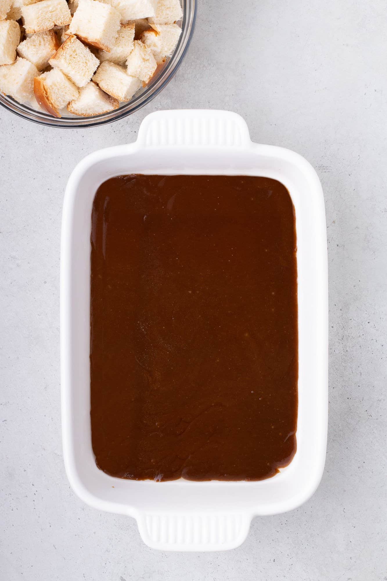 A white baking dish filled with a dark brown liquid mixture sits on a light surface. A bowl containing cubed bread pieces is visible above it.