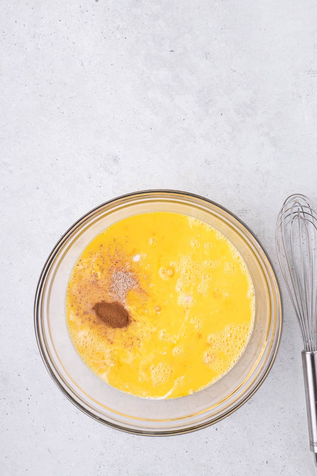 A glass bowl with beaten eggs and spices next to a metal whisk on a gray countertop.