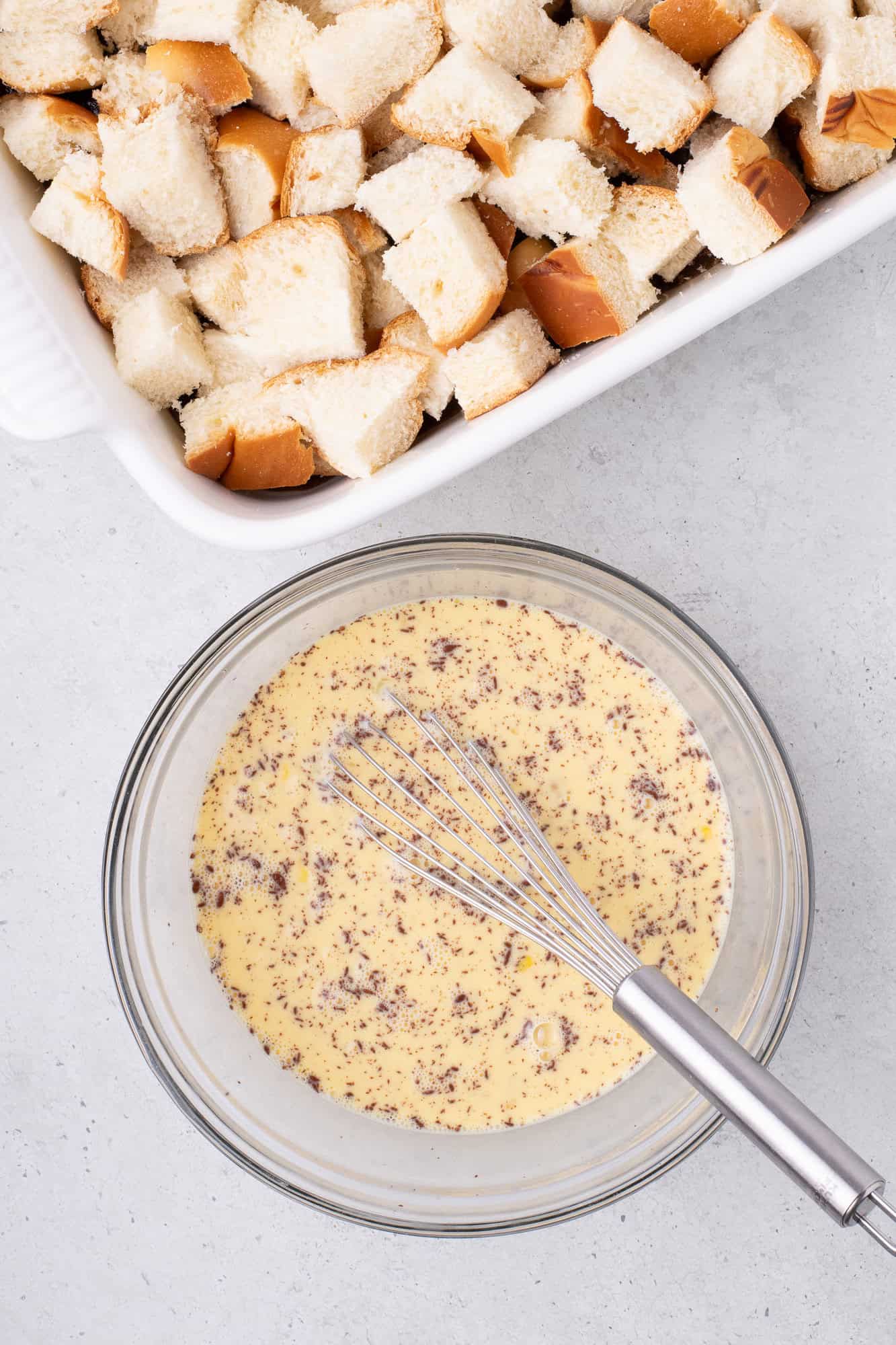 Cubed bread in a white baking dish; a glass bowl with a whisk and a speckled egg mixture beside it on a gray surface.