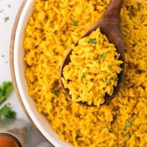 A wooden spoon holds a serving of yellow rice garnished with herbs over a bowl filled with the same rice.