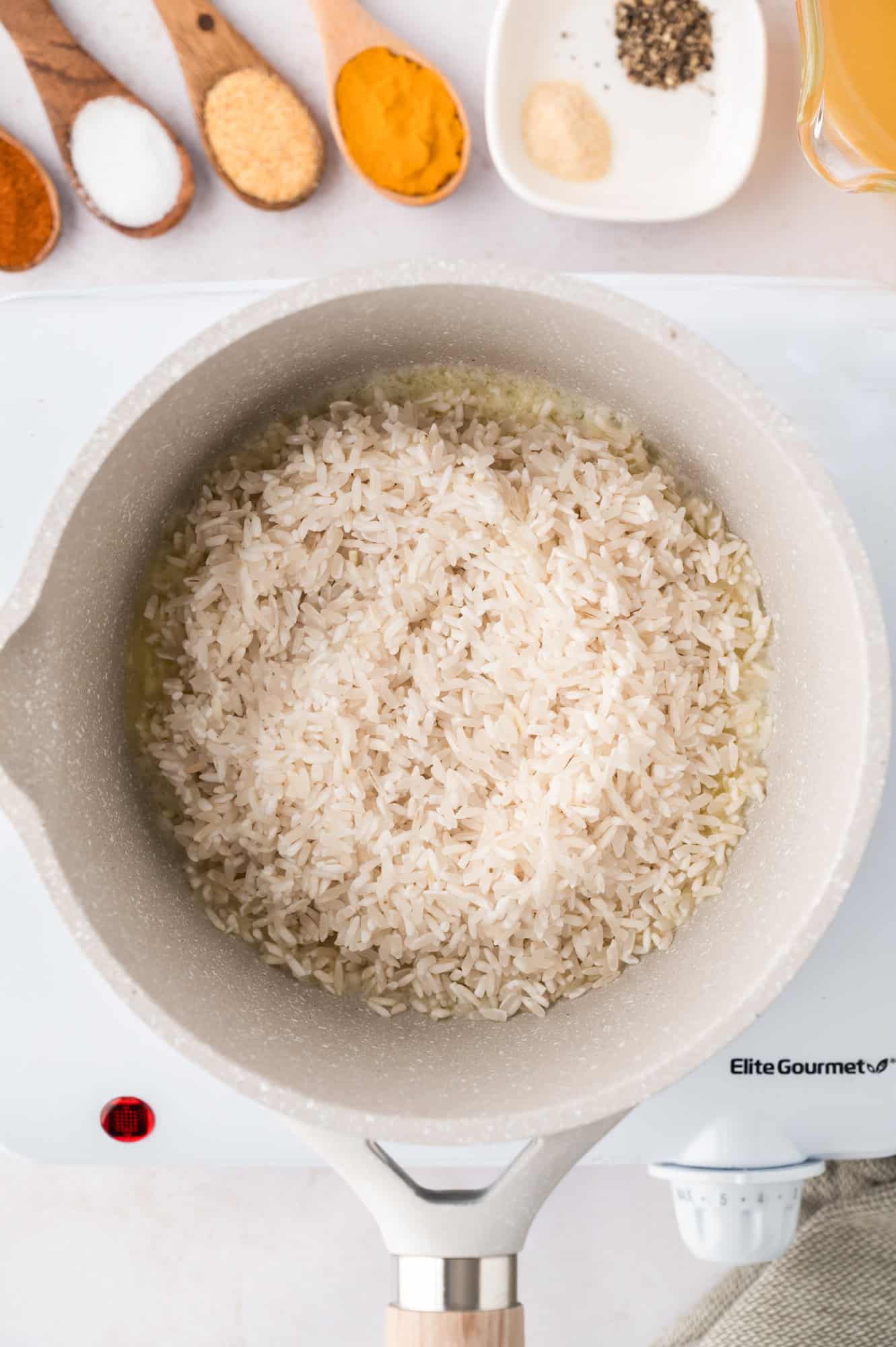 A pot of uncooked white rice sits on a stove. Various spices and ingredients are visible in small bowls nearby.