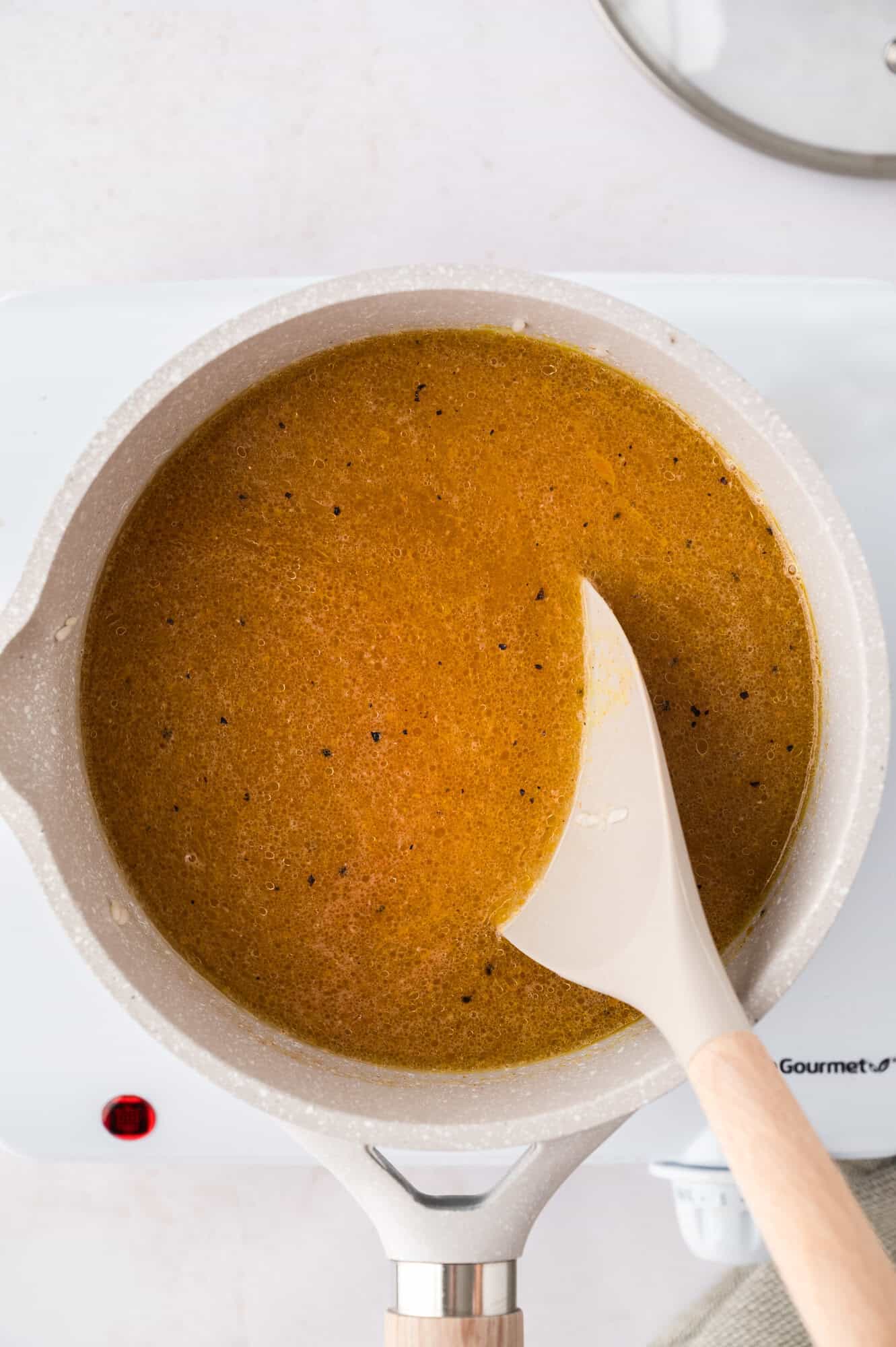 A pot on a stove contains a brown liquid being stirred with a white spatula.