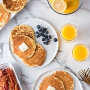 A plate of pancakes with butter and blueberries, bacon, a bowl of lemons, and two glasses of orange juice on a marble surface.