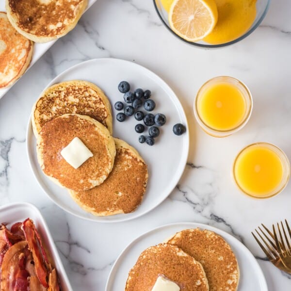 A plate of pancakes with butter and blueberries, bacon, a bowl of lemons, and two glasses of orange juice on a marble surface.