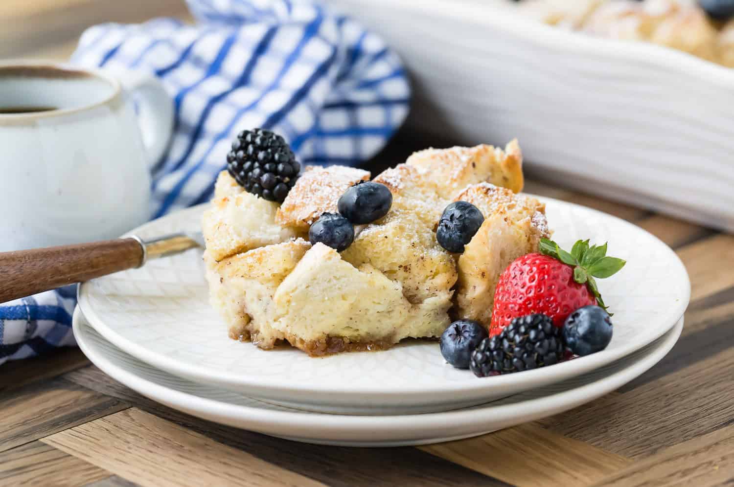 A serving of bread pudding topped with powdered sugar, blueberries, blackberries, and a strawberry, on a white plate with a fork. 