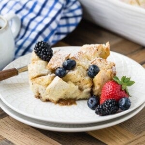 A slice of bread pudding with scattered blackberries and blueberries, alongside a strawberry, on a white plate.