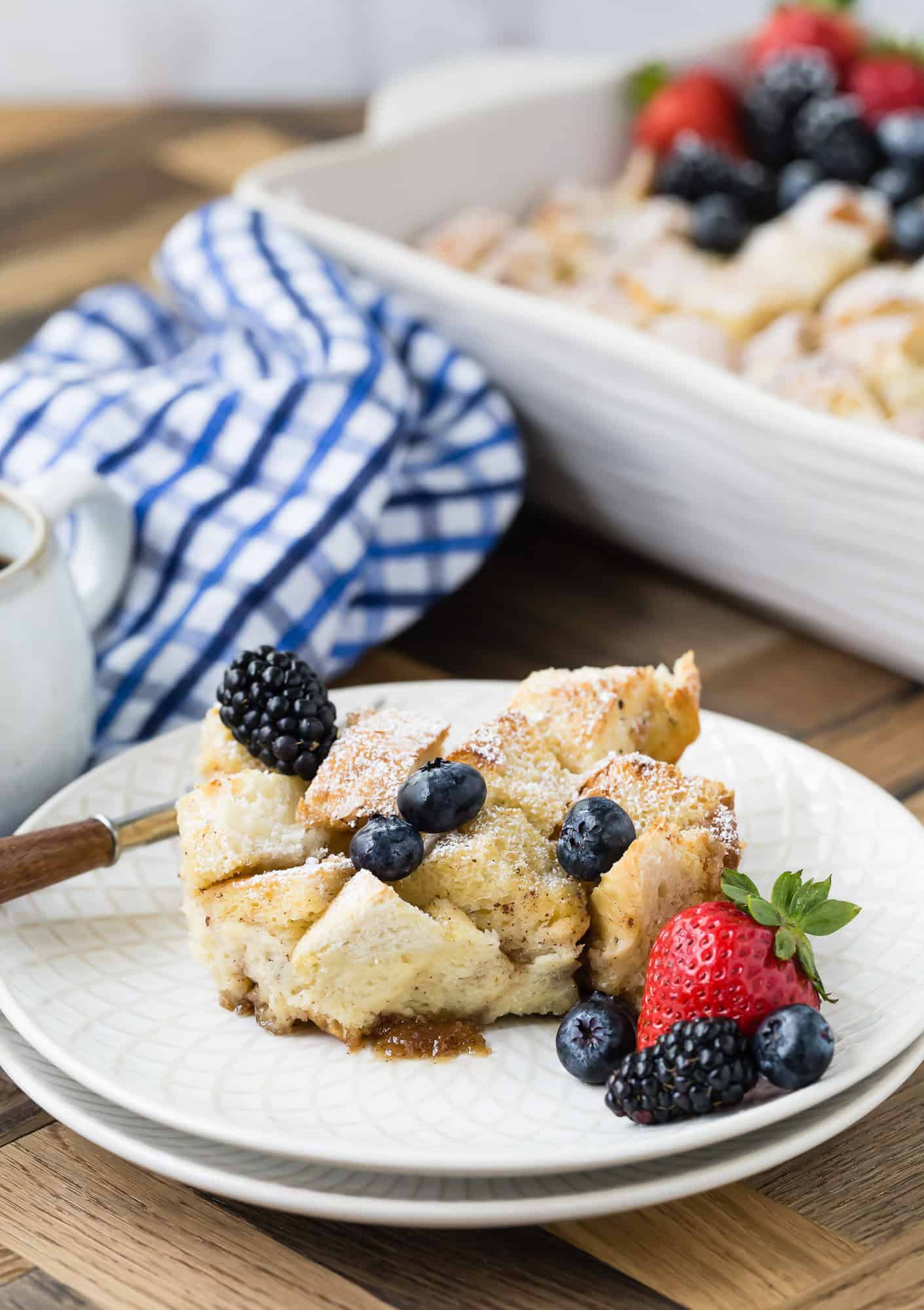 A slice of bread pudding on a plate, topped with blackberries and blueberries. A casserole dish and a blue checkered cloth are in the background.