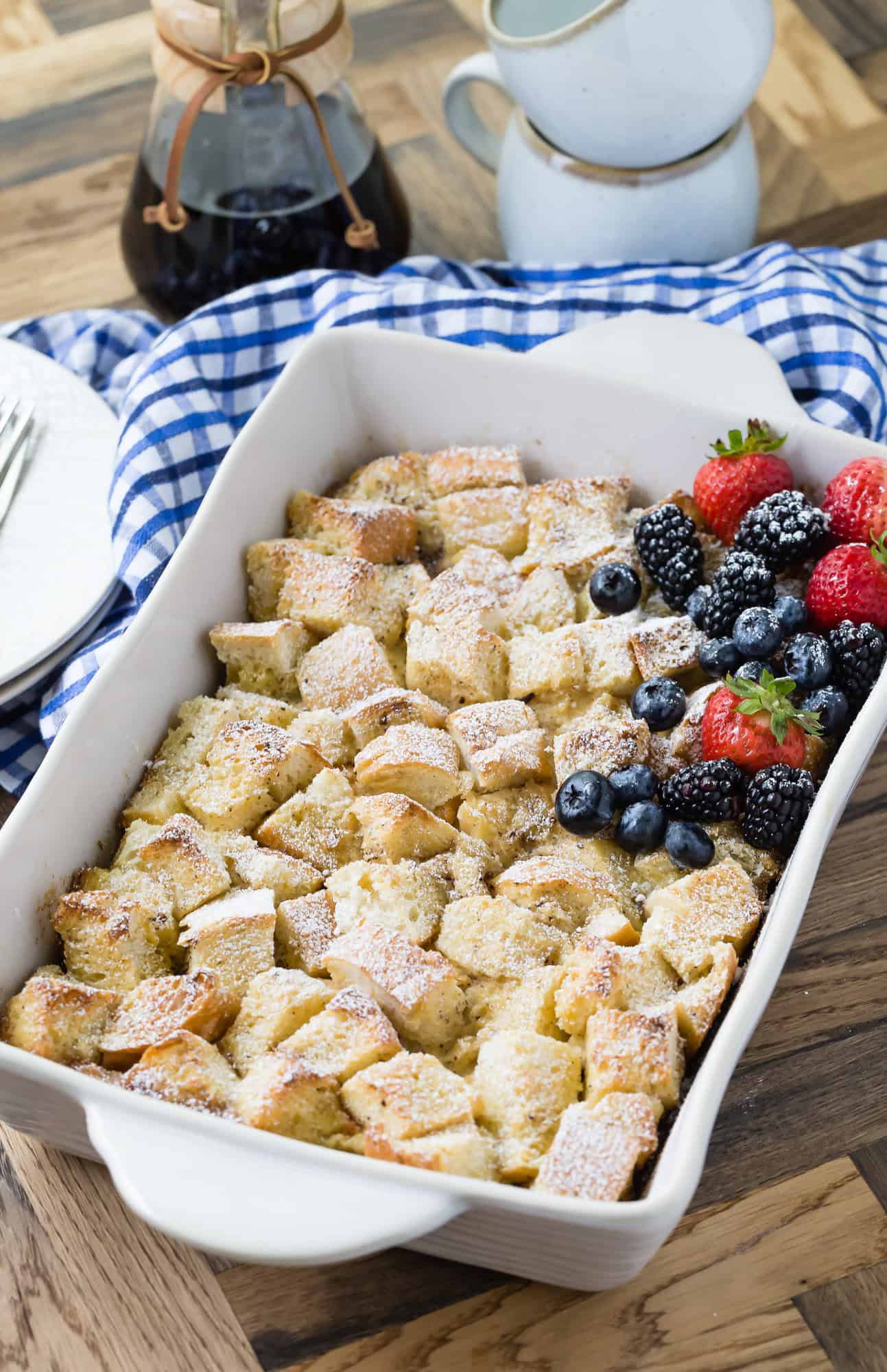 Baked French toast casserole with powdered sugar, strawberries, blueberries, and blackberries in a white dish on a wooden table with a blue checkered cloth.