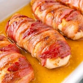 Bacon wrapped chicken breasts in a dish, cooked to a golden brown and surrounded by a layer of juices.