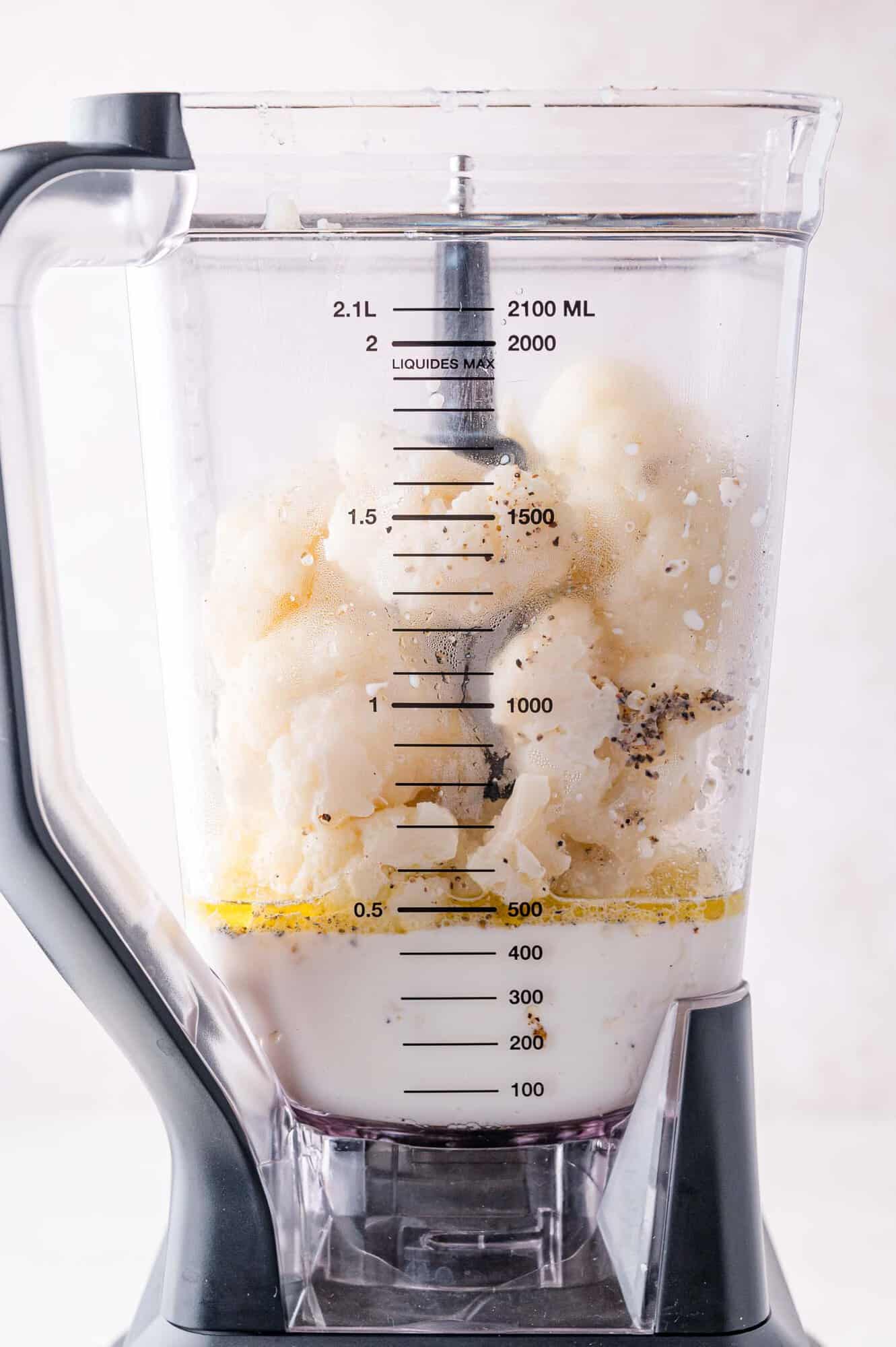 A blender filled with cauliflower, milk, herbs, and black pepper on a white surface.