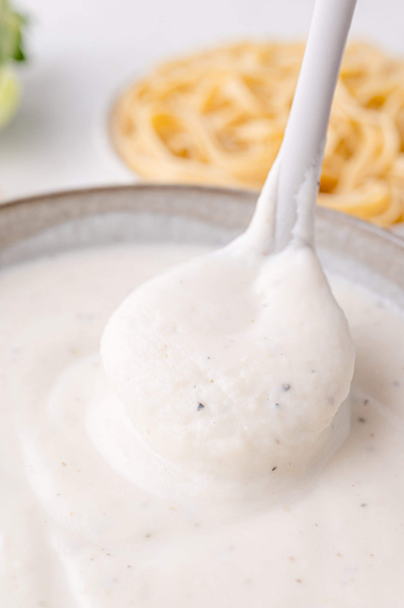A ladle scoops creamy white Alfredo sauce from a bowl, with some fettuccine pasta visible in the background.