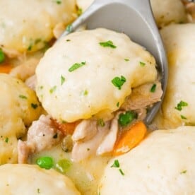 A close-up of chicken and dumplings in a creamy sauce with carrots and peas. A spoon lifts one dumpling, topped with chopped herbs.