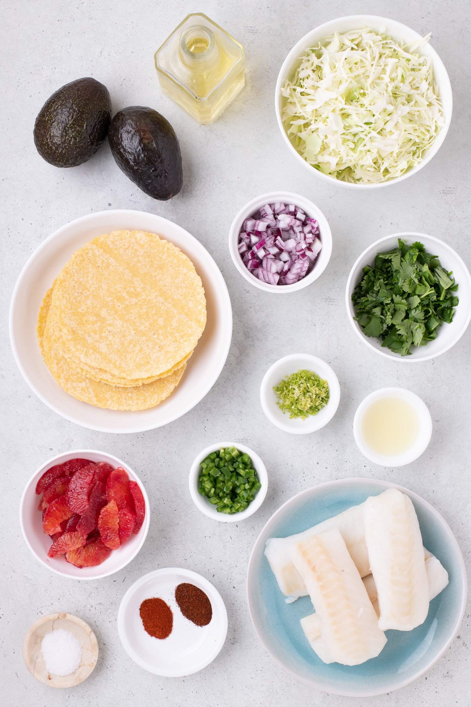 Ingredients for fish tacos laid out: corn tortillas, fish fillets, sliced cabbage, avocado, lime zest, lime juice, red onion, cilantro, jalapeño, chili powder, and salt.