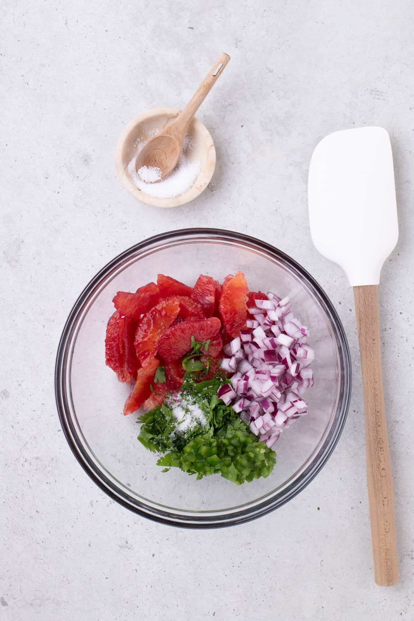 Glass bowl with diced tomatoes, red onion, jalapeño, and cilantro. Nearby are a small wooden bowl with salt and a white spatula on a gray surface.