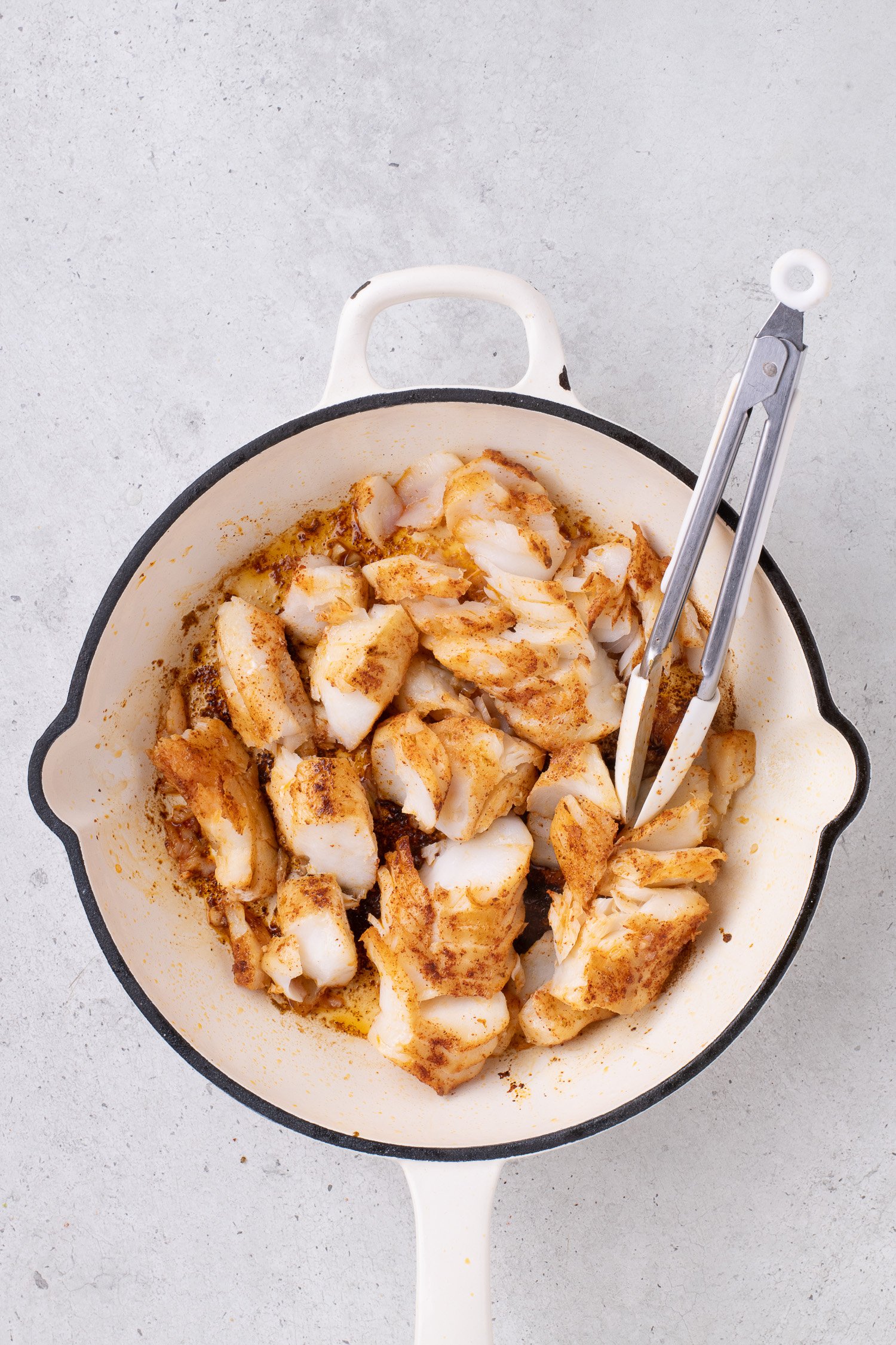 Cooked pieces of seasoned fish in a white skillet with metal tongs on a light surface.