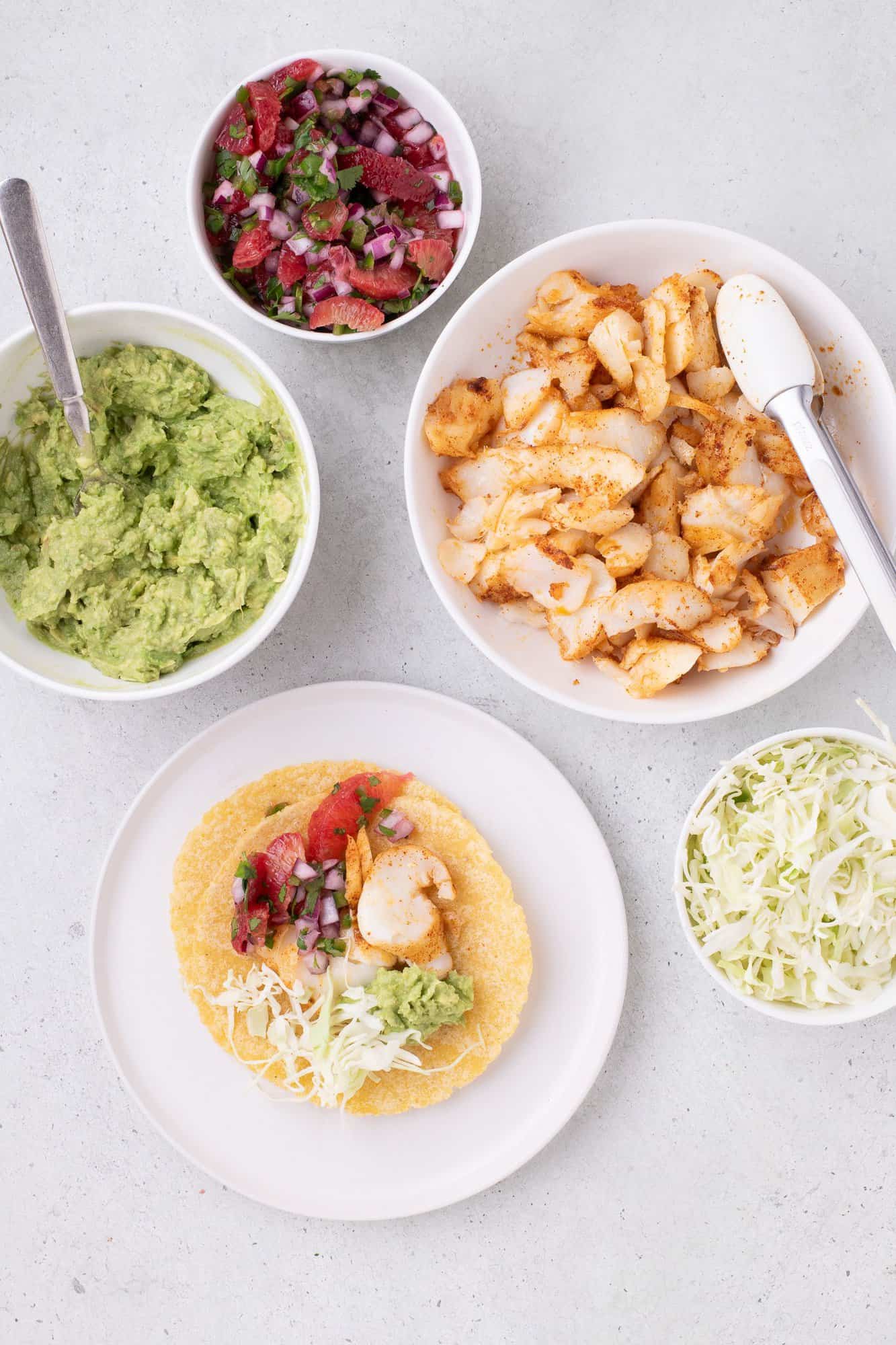 Taco ingredients with corn tortillas, seasoned fish, guacamole, cabbage, and salsa displayed on a light surface.