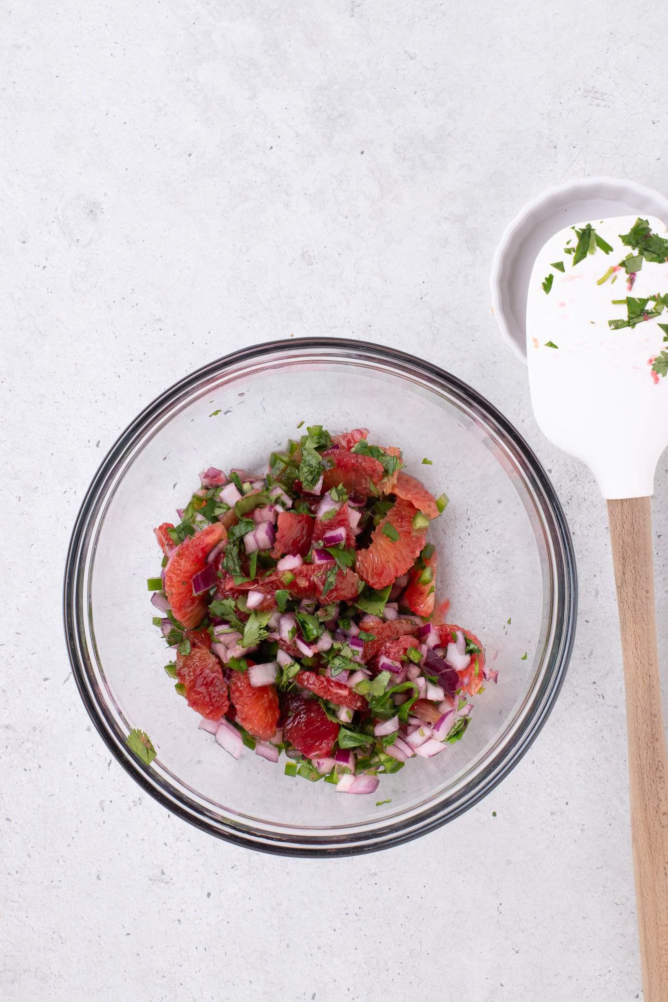 A bowl of diced red onions, cilantro, and grapefruit segments, with a spatula nearby on a light-colored surface.