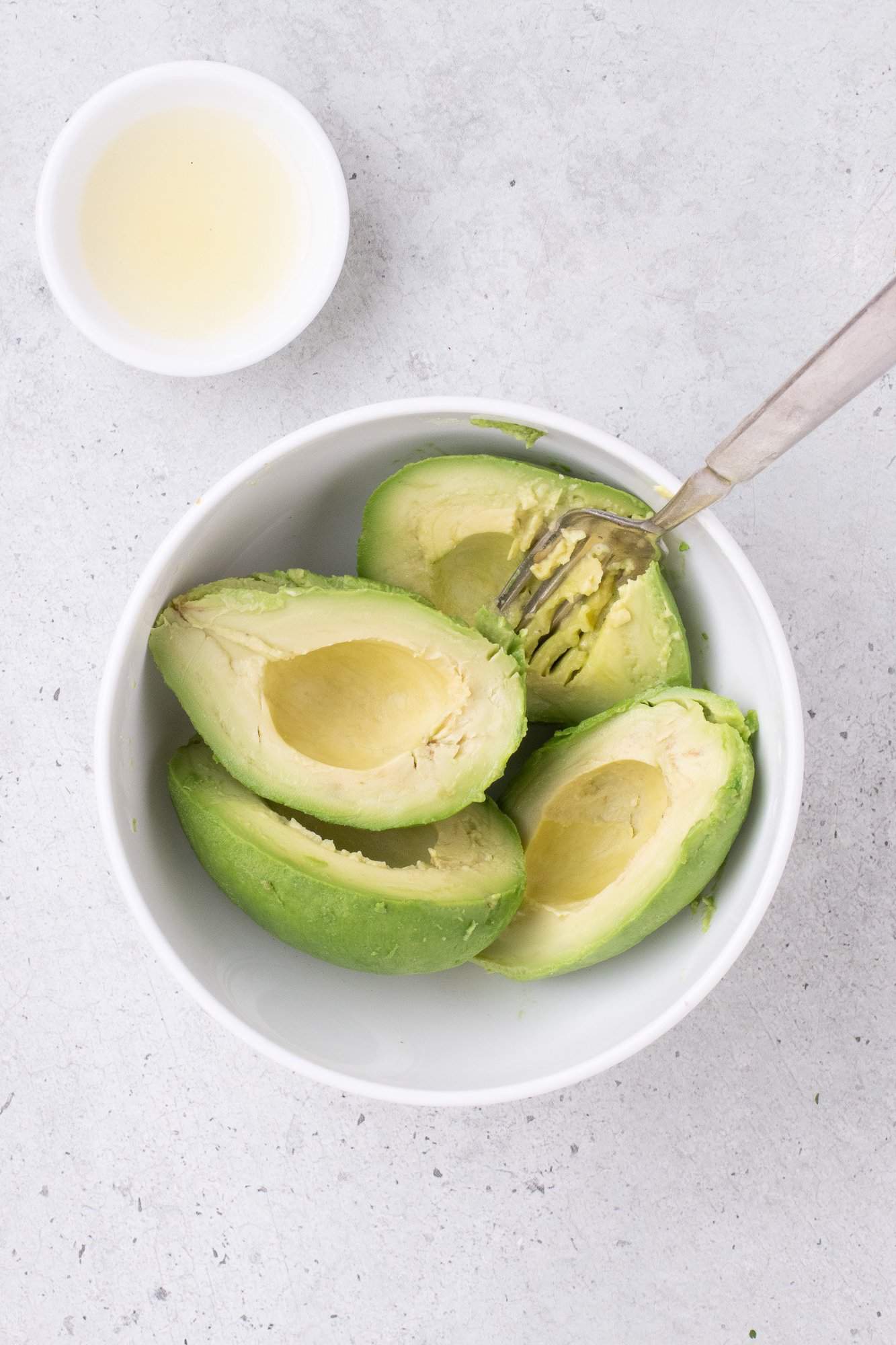 A bowl with four avocado halves and a fork next to a small bowl of lemon juice on a light gray surface.