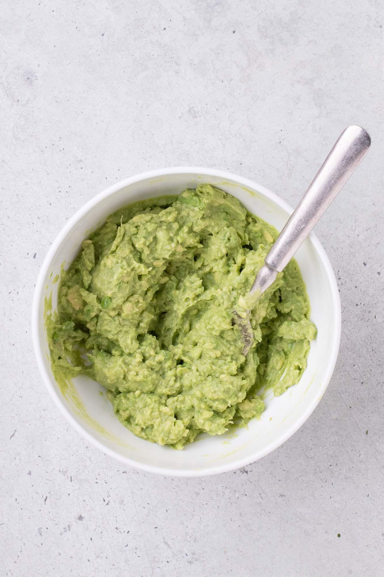 Bowl of mashed guacamole with a spoon on a light gray surface.