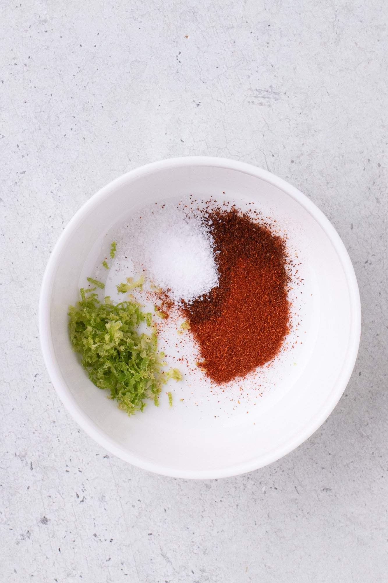 A bowl with salt, paprika, and lime zest on a light textured surface.