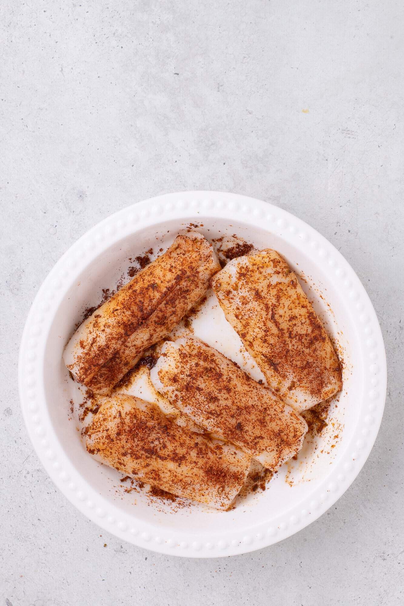 Four raw fish fillets in a white dish, sprinkled with seasoning on a light gray background.