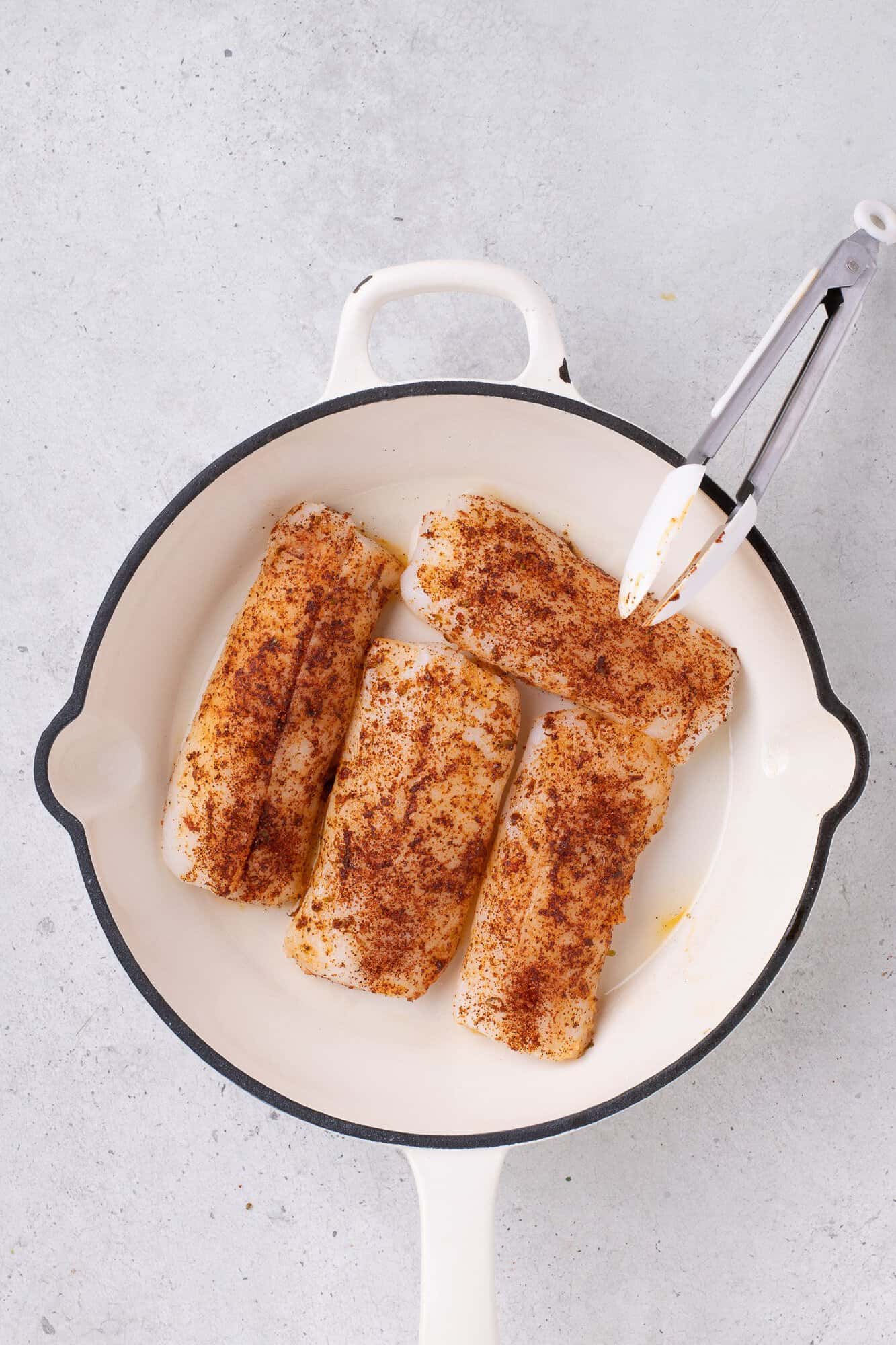 Four seasoned fish fillets in a white frying pan with tongs, placed on a light gray surface.