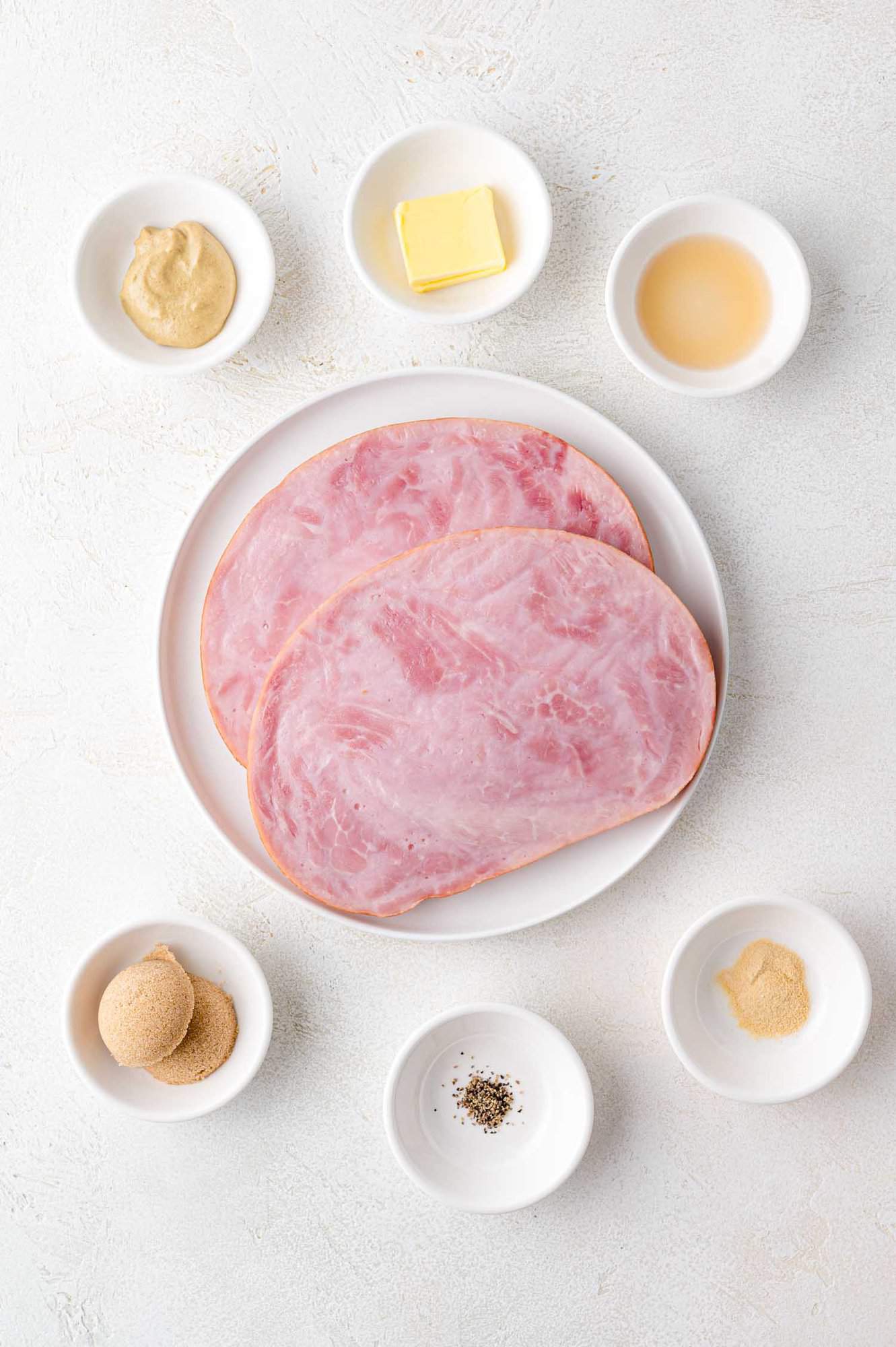 Plate with two slices of ham surrounded by small bowls containing mustard, butter, vinegar, brown sugar, black pepper, and powdered spice on a light textured surface.