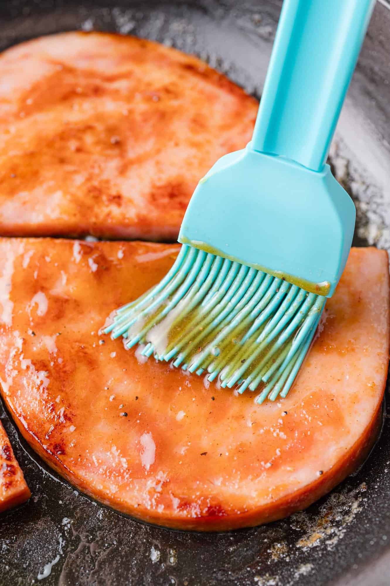 Two slices of ham being glazed with a blue brush in a frying pan.