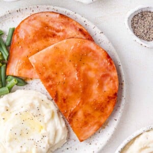 Two slices of glazed ham on a plate with mashed potatoes and green beans, seasoned with pepper.